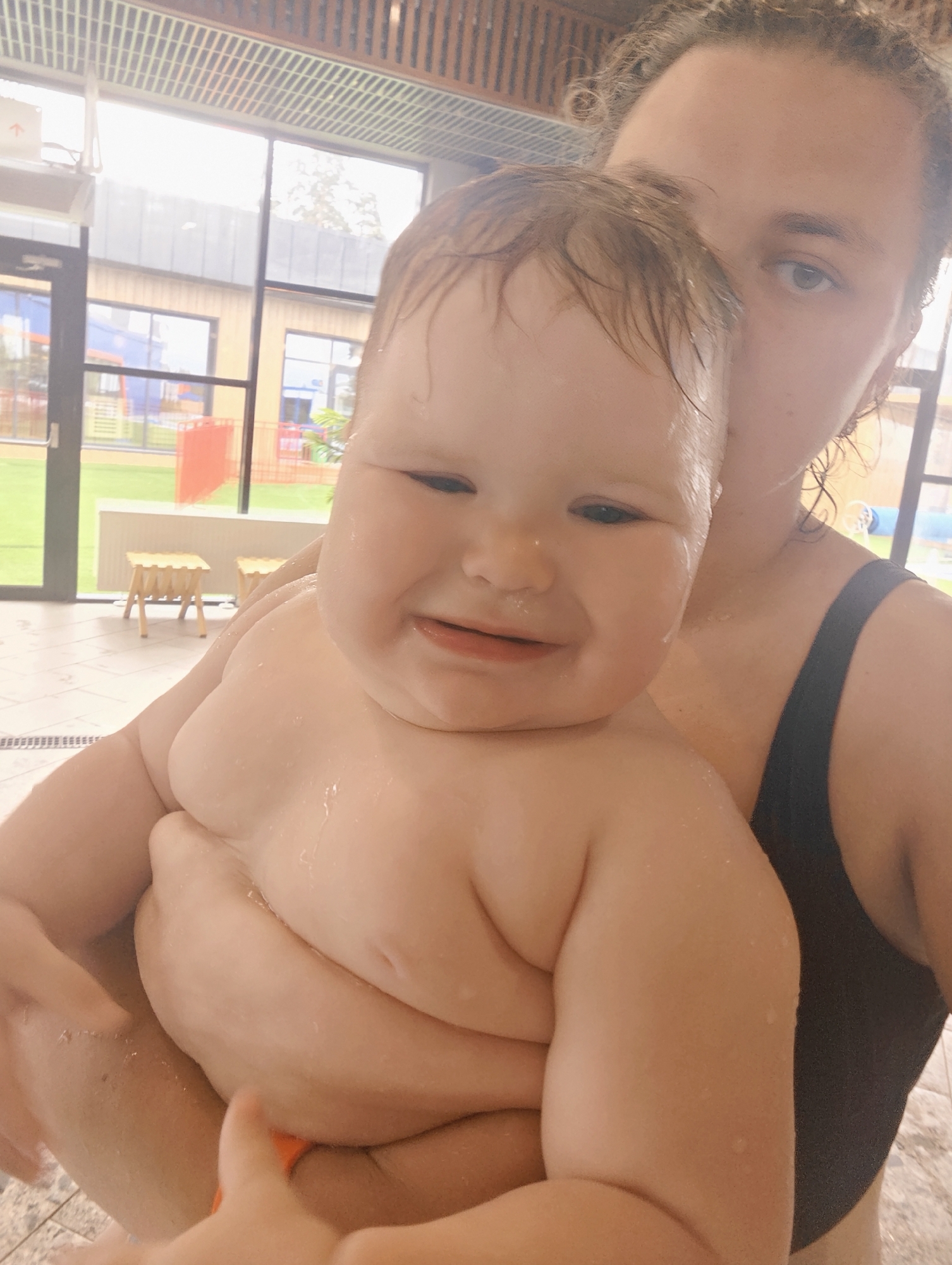 A woman is holding a baby indoors, seemingly in a swimming pool area. The woman is wearing a black bathing suit. Both the woman and baby have wet hair and skin. The background features large windows, a wooden-paneled ceiling, and tile flooring characteristic of an indoor pool environment. The location appears to be a modern swimming complex. While the image suggests an indoor pool scene, there's nothing in the image to specifically indicate that it is located in Votkinsk, Russia. Therefore, the location cannot be confirmed.