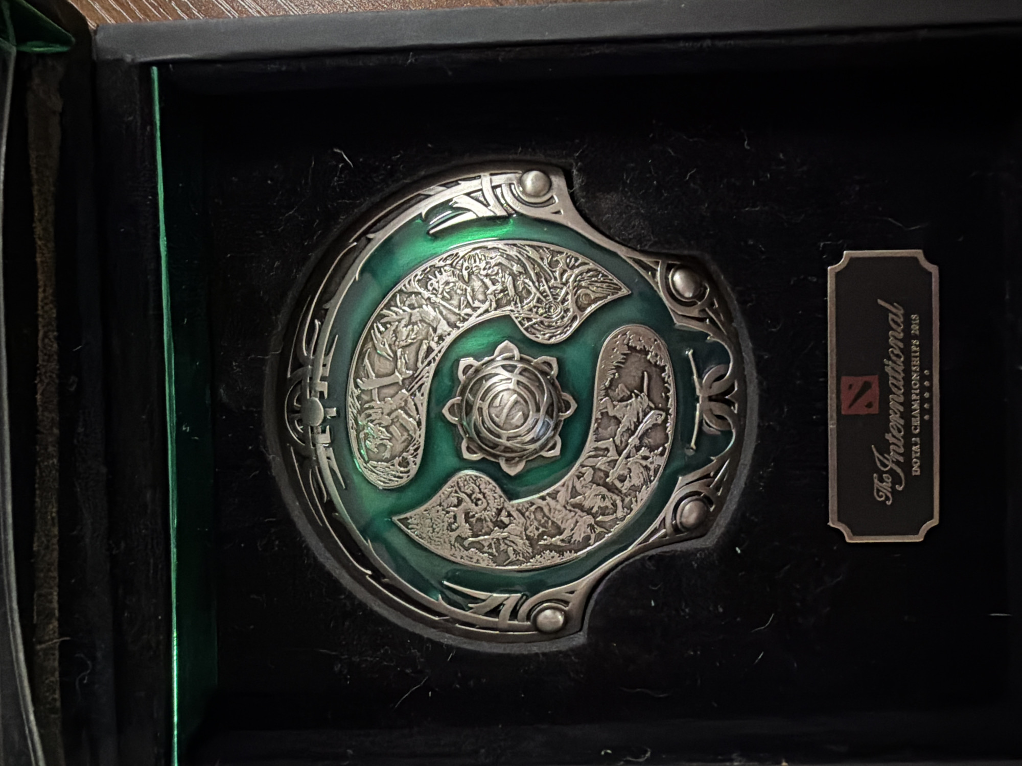 The image displays a close-up, overhead view of a round, metallic medallion with intricate engravings and green enamel accents, resting in a black, velvet-lined box. The medallion features a central circular design with radiating patterns, surrounded by a larger circular motif with depictions of various creatures or figures. The outer edge of the medallion is ornamented with decorative metalwork. Below the medallion, a rectangular plaque with rounded corners is affixed to the box's lining. The plaque has text that reads "The International" in a stylized font, followed by "DOTA 2 CHAMPIONSHIPS 2018" and a series of dots or stars. The box itself appears to be made of dark material, possibly wood or reinforced cardboard, with a dark green accent line visible along the top edge of the opened lid. The overall presentation suggests a trophy or commemorative item. The location is provided as Yablonovskiy, Russia.