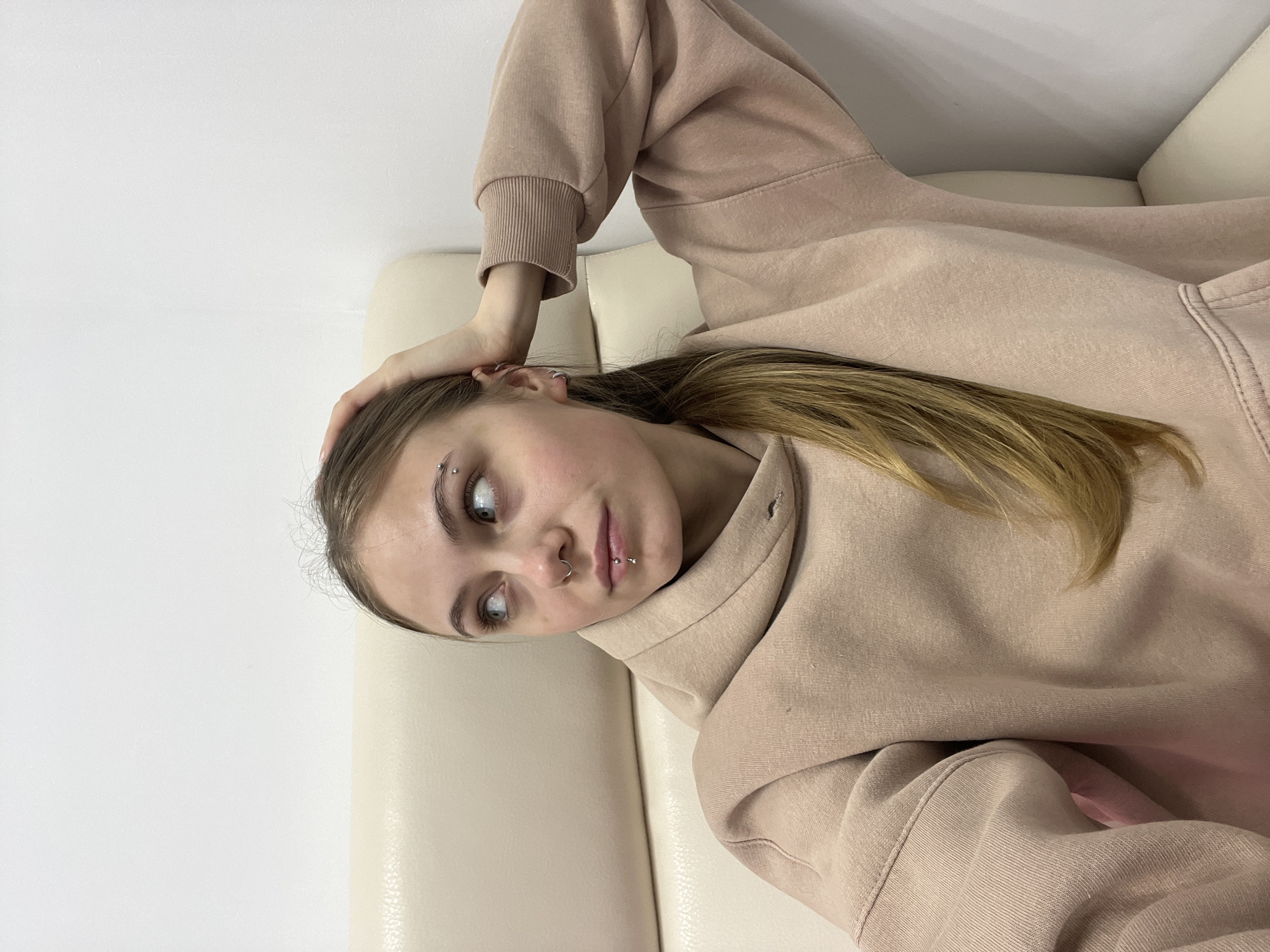 The image captures a young woman in a casual, indoor setting, seen from a slightly high angle and rotated 90 degrees counter-clockwise. She is lying down, with her head resting on her left hand, which is bent at the elbow. Her light brown or blonde hair, styled to fall over her shoulder, is visible on the right side of the frame.
The woman is wearing a light beige or peach-colored hoodie, which covers most of her upper body and arms. Her left arm is visible, with her hand supporting her head, while her right arm is bent with the sleeve of the hoodie covering it. The fabric of the hoodie shows some texture and subtle folds, particularly around the sleeves and torso.
Her face is angled towards the camera, and she is looking slightly upwards and to the right with light-colored eyes, possibly blue or grey. She has several noticeable piercings: two silver barbells on her left eyebrow, a silver hoop in her septum, and a silver labret piercing below her lower lip. A small silver hoop earring is also visible on her left ear. Her expression appears calm and contemplative, or perhaps slightly quizzical.
The background consists of a plain white wall on the left and upper left, and a light-colored, possibly beige or cream, upholstered surface on the right and underneath her, suggesting she is lying on a sofa, bed, or similar piece of furniture. The lighting is soft and even, indicating an indoor environment without harsh shadows. There is no visible text in the image.