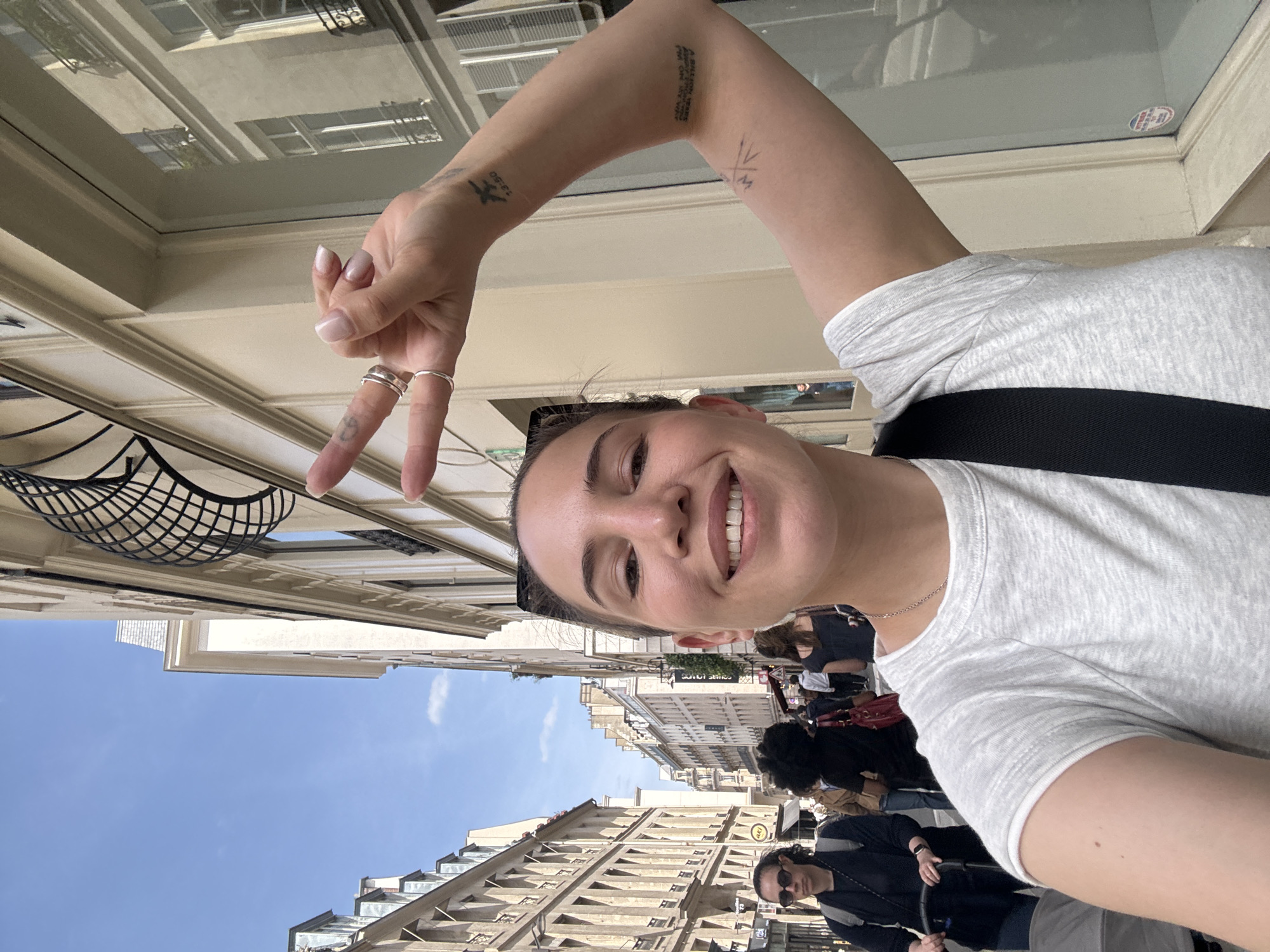 The image is a bright, upright selfie of a young woman, taken outdoors on a sunny day in a bustling city street identified as Paris, France. The woman is smiling broadly at the camera, revealing her teeth, and appears cheerful. She is wearing a light gray short-sleeved t-shirt and a dark strap, possibly from a bag or camera, across her shoulder.
Her left arm is raised towards the camera, with her hand forming a peace sign. On her left ring finger, she wears two simple silver rings. Her fingernails are neatly manicured with a light, natural polish. Several tattoos are visible on her arm: on her inner forearm, the French phrase "A BIENTOT MON AMOUR" (meaning "See you soon my love"); near her wrist, a small script that looks like "12.50" along with a small bird outline; and higher up her arm, a minimalist geometric design.
The background features typical Parisian architecture, with multi-story stone buildings lining a street under a clear, bright blue sky with a few wispy clouds. Windows, balconies, and decorative elements are visible on the buildings. Several other people can be seen walking or congregating in the background, suggesting a lively urban setting. Some visible text includes a green sign on a distant building, possibly displaying "LUXURY JEWELRY," and a yellow sign with faintly visible letters that might spell "L'EST." The time of day is clearly daylight.