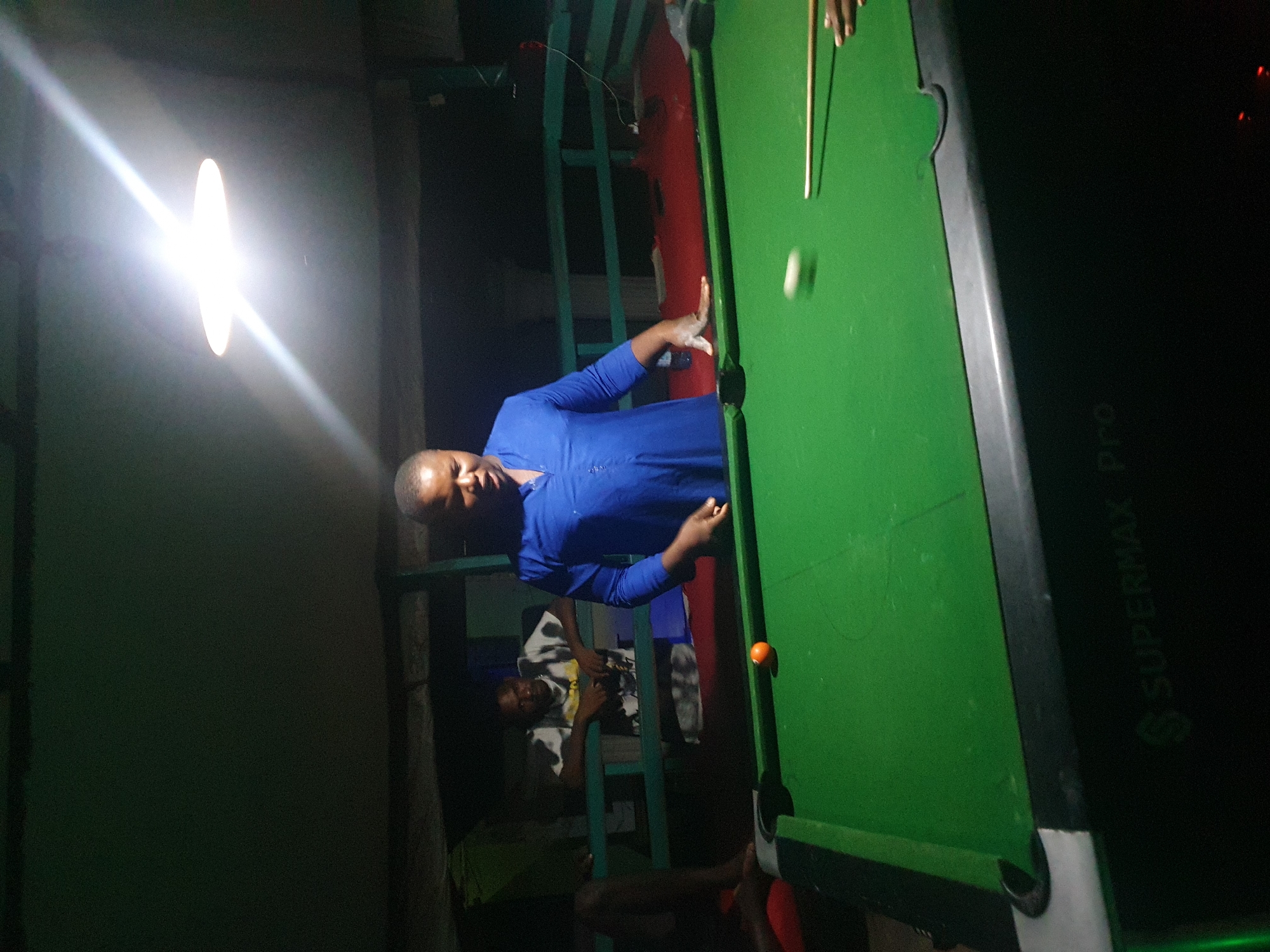 In Lokoja, Nigeria, a group of individuals is gathered around a billiard table under dim lighting, likely at a local establishment. The scene captures a moment of leisure, with one person actively engaged in a game of pool. The lighting, originating from an overhead fixture, creates a bright, starburst effect and illuminates the pool table, highlighting the green felt and the balls.
The focal point is a person wearing a blue shirt, positioned to the right of the frame, with their hand extended towards the billiard cue. They appear to be either striking the cue ball or in the process of preparing to do so. A cue ball is visible in motion, blurred by its speed, indicating an active game. To the left, a single orange billiard ball rests on the felt. Another person, wearing a white and black patterned shirt, leans on a railing in the background, observing the game. The setting seems to be an open-air or semi-open space, with a railing separating the billiard area from the surroundings. The overall atmosphere is informal and suggests a recreational gathering.