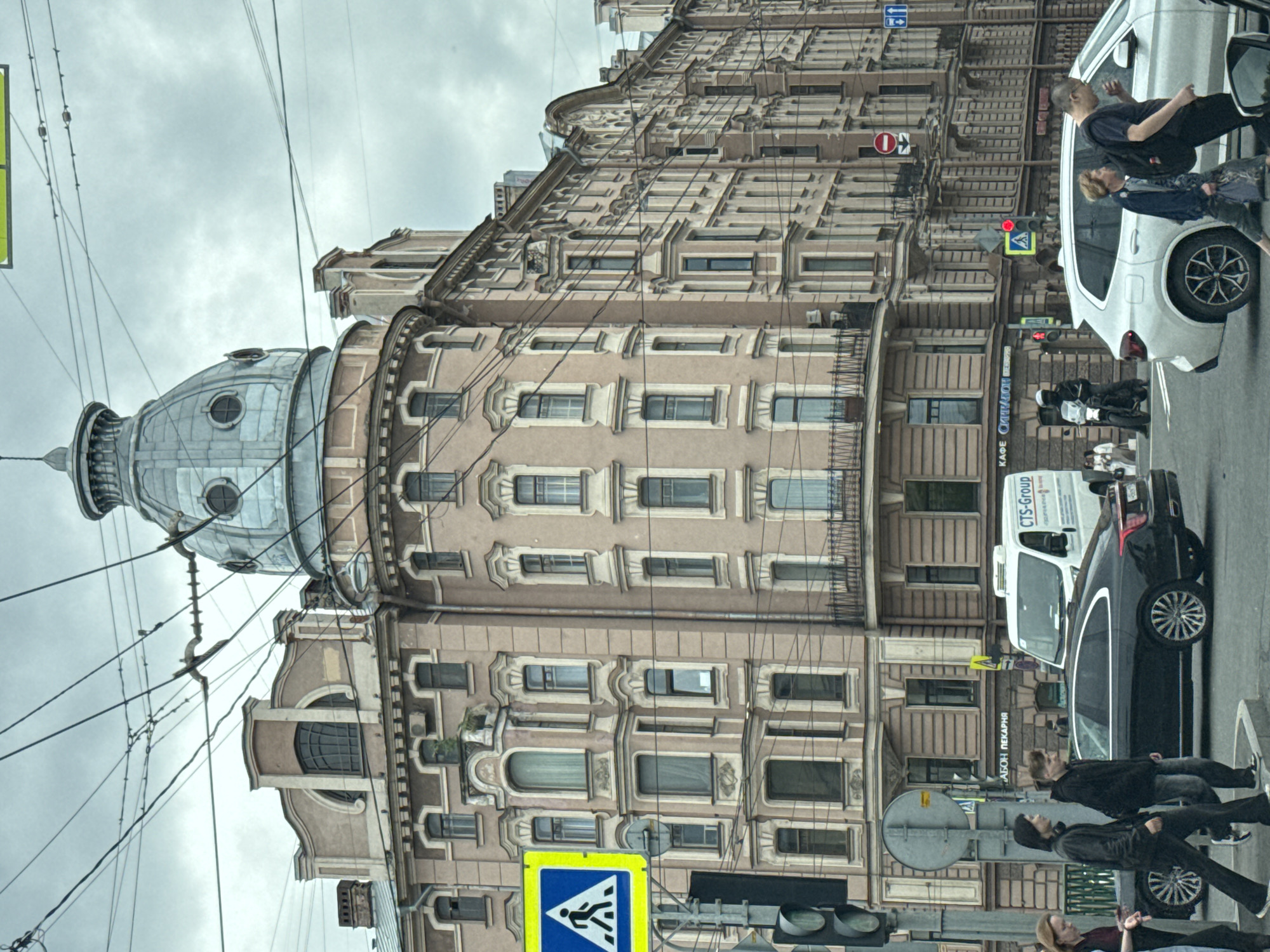 The image captures a view of a street in Saint Petersburg, Russia, on a cloudy day. The scene is dominated by a grand, historic building with a distinctive dome and ornate facade. In the foreground, there is a busy street with several vehicles, including a black sedan, a white SUV, and a white van. Pedestrians are visible crossing the street, and traffic lights and street signs are present. The architecture suggests a European city, and the presence of Russian text on a sign confirms the location as Saint Petersburg. The overall atmosphere is one of urban activity, with elements of historical architecture blending with modern city life.