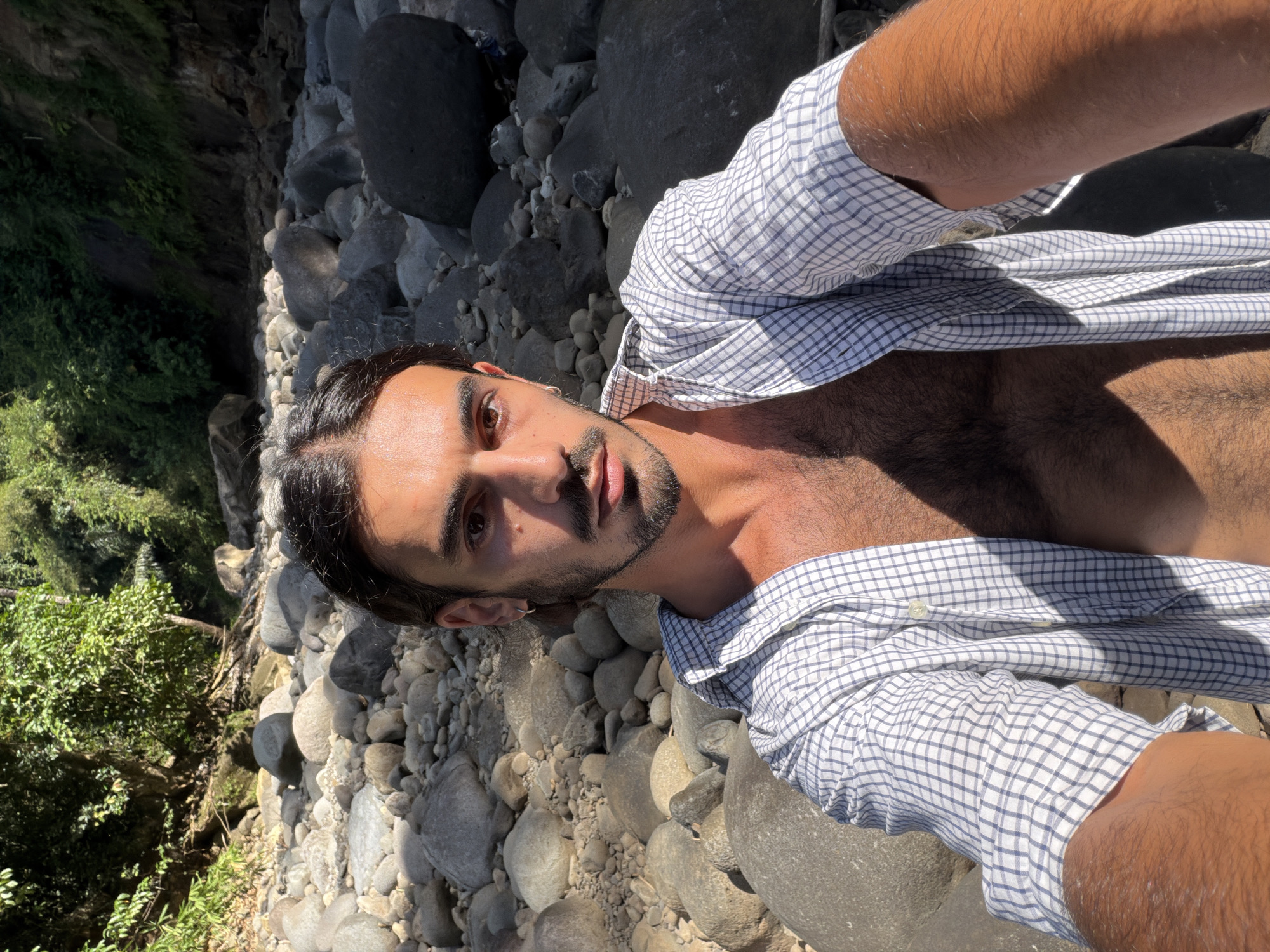 A selfie captures a man with dark hair and a beard, wearing an open, checkered shirt revealing his chest. He is positioned on a rocky terrain, likely a riverbank or stream bed, evidenced by the smooth, rounded rocks. Lush vegetation is visible on the left side of the frame, suggesting a natural environment. The lighting indicates it is daytime with clear skies.
The location is the city of Selemadeg Timur, Indonesia.