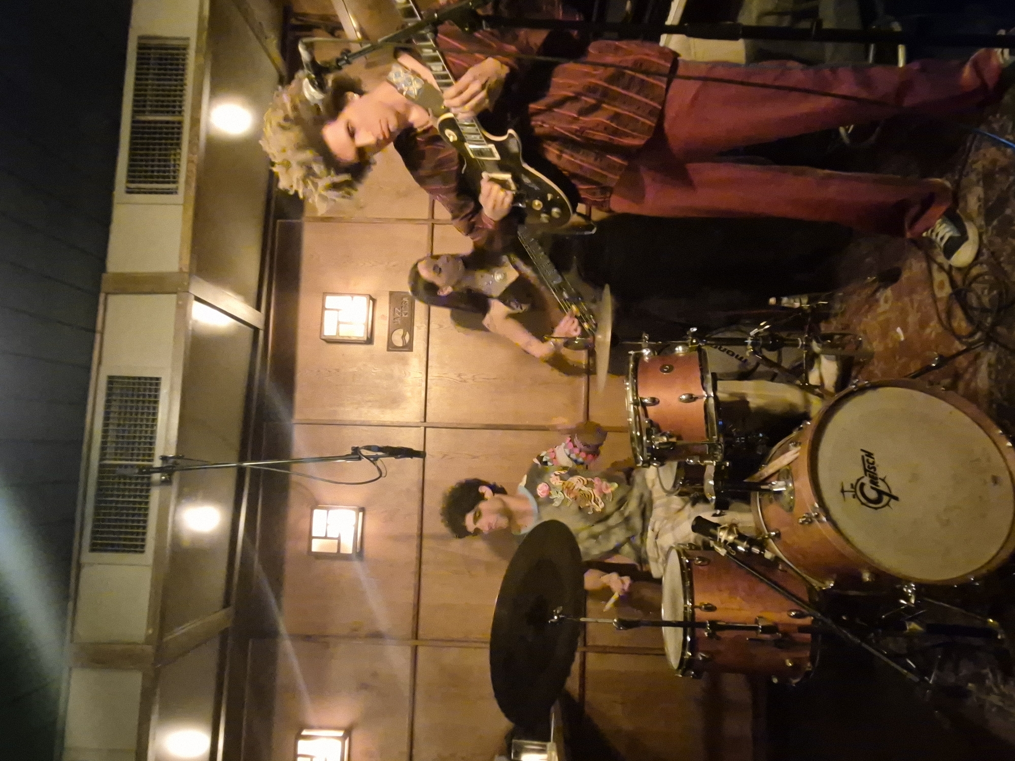 The image shows a musical band performing on a stage inside a venue. There are three band members visible. One man, wearing a burgundy striped suit, is playing an electric guitar. Another woman is playing a guitar as well. The third man is seated behind a drum kit with a “Gretsch” logo on the bass drum, playing the drums. The interior walls are wood-paneled with several square light fixtures. A ventilation grill is visible near the upper left of the image. Based on an unconfirmed logo on the wall behind the band, the image may have been taken at Jazz Kissa, a jazz club located in Tel Aviv, Israel.