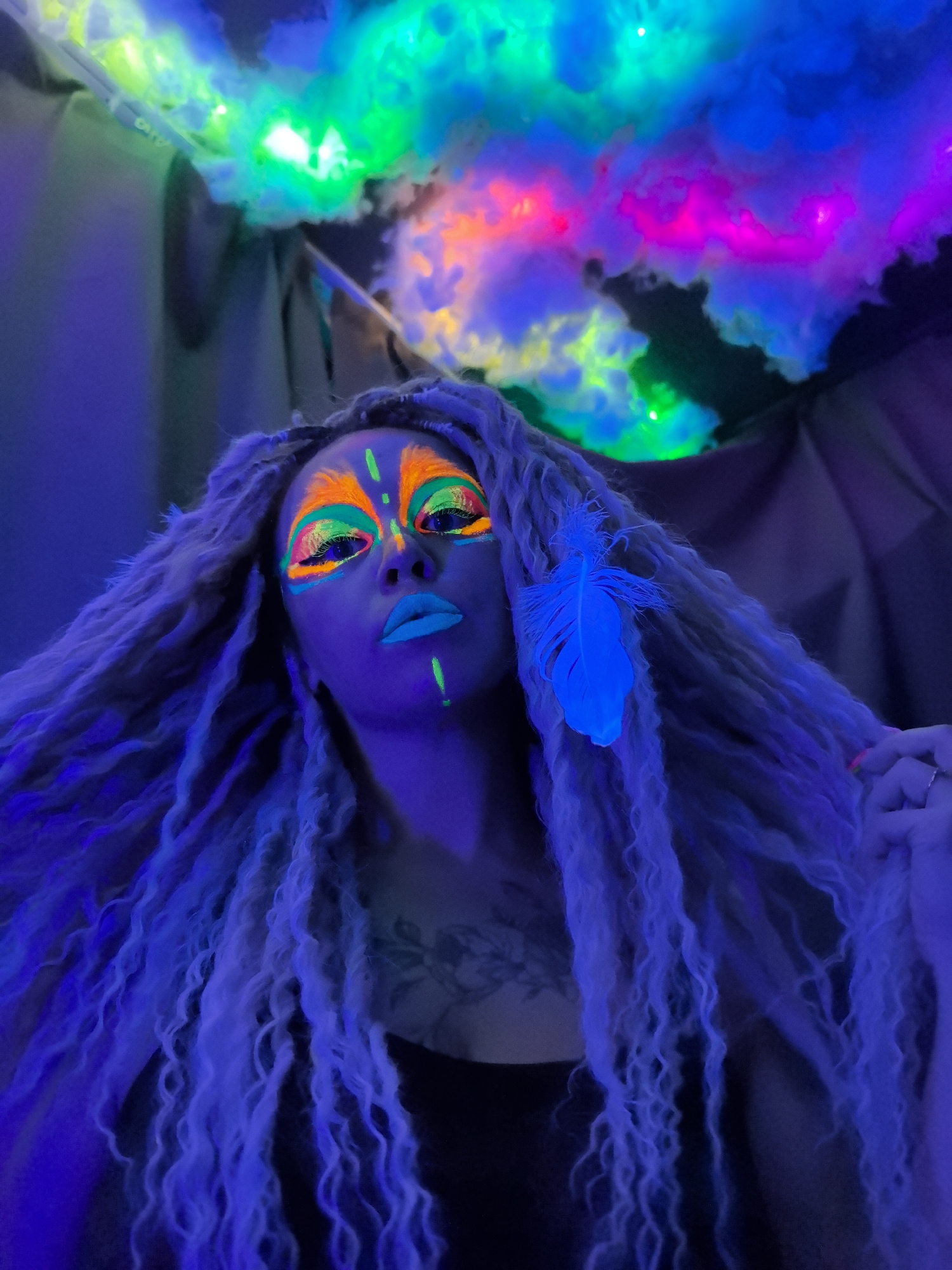 A woman is depicted in a low-angle close-up shot, exhibiting fluorescent makeup and accessories under a blacklight. Her makeup consists of orange lines around her eyes and extending upwards onto her forehead, accented by green lines; she has blue lips and a green line extending down from her lower lip. Her hair is long and wavy and adorned with a blue feather. Above her, simulated clouds are illuminated by a multicolored light display. A tattoo is visible on her chest. The background consists of dark curtains or walls. There is no indication of this being in the city of Noviy, Russia.