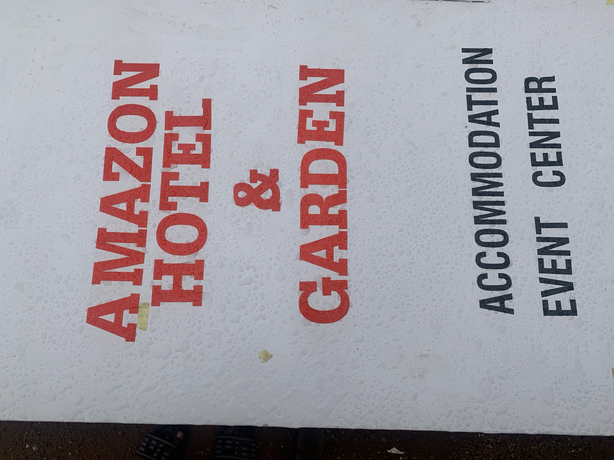The image shows a white rectangular sign, oriented vertically, covered in numerous small water droplets, suggesting it is outdoors and has been exposed to moisture. The sign is positioned above a dark, granular surface, possibly ground or pavement, with parts of a person's feet, clad in dark open-toed sandals, visible at the very bottom edge.
The sign features text in two distinct sections. On the left, prominent red, sans-serif, uppercase lettering spells out "AMAZON HOTEL & GARDEN." Small, yellowish pieces of tape or adhesive are visible on some of the red letters, specifically on the 'A' of "AMAZON" and near the '&' symbol.
To the right, smaller, black, sans-serif, uppercase text is vertically stacked. The top line reads "ACCOMMODATION," and the bottom line reads "EVENT CENTER."
The sign appears to be advertising a hospitality business. Given the context of "New Stadium, Nigeria," this sign likely indicates the presence of a hotel, garden, accommodation facilities, and an event center in the vicinity of a stadium. The water droplets suggest recent wet weather conditions.