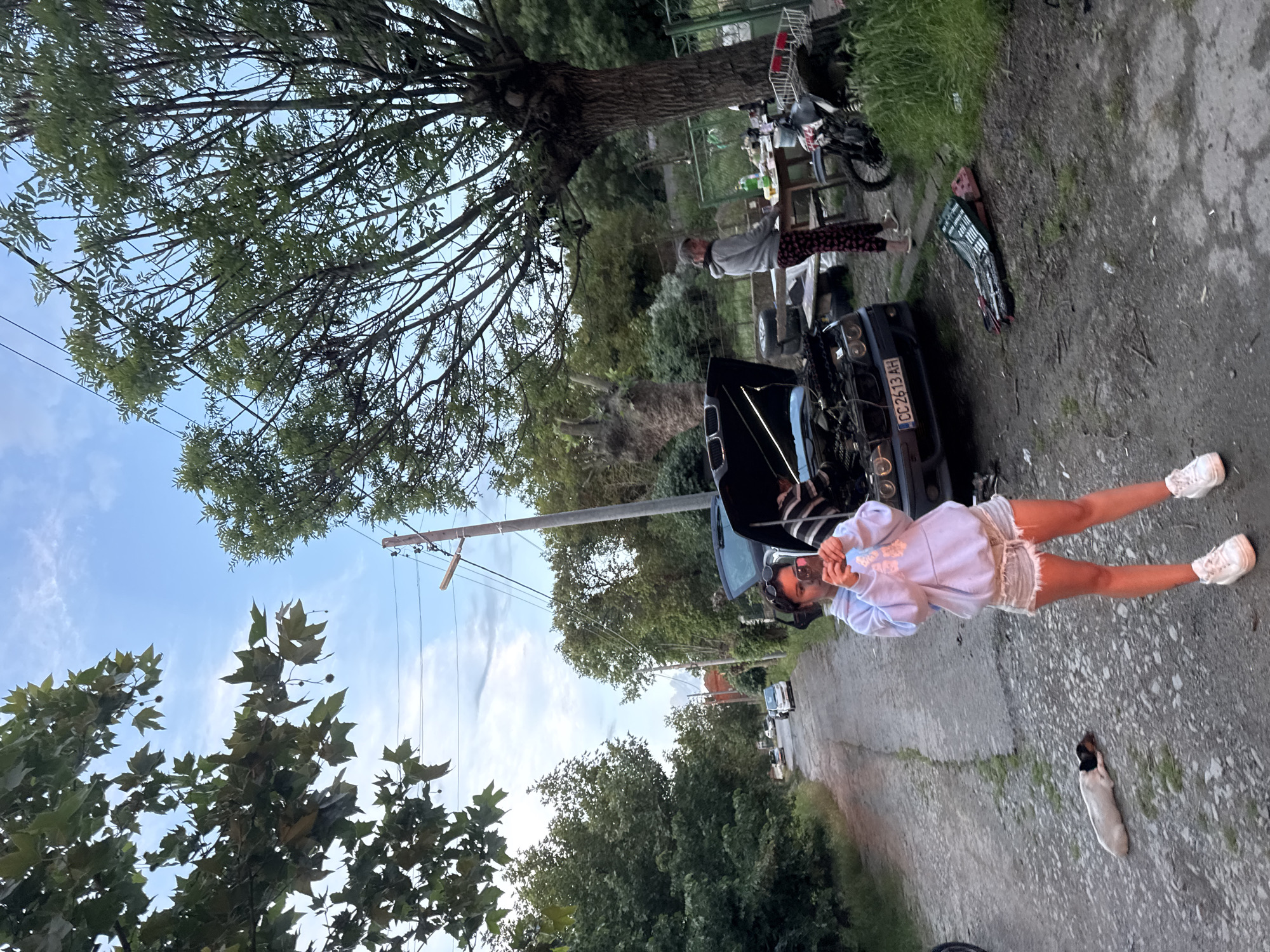 The image appears to be taken in Ruzhitsa, Bulgaria, based on the license plate "CC 2613 AH" visible on the black car. A young woman wearing a blue sweater and shorts stands in front of the car with its hood open. To the right, a white and brown dog lies on the ground. In the background, there is a motorcycle, table with items, and a person wearing gray sweatpants and a sweater. The setting appears to be a rural street with trees and power lines visible. The sky is partly cloudy.