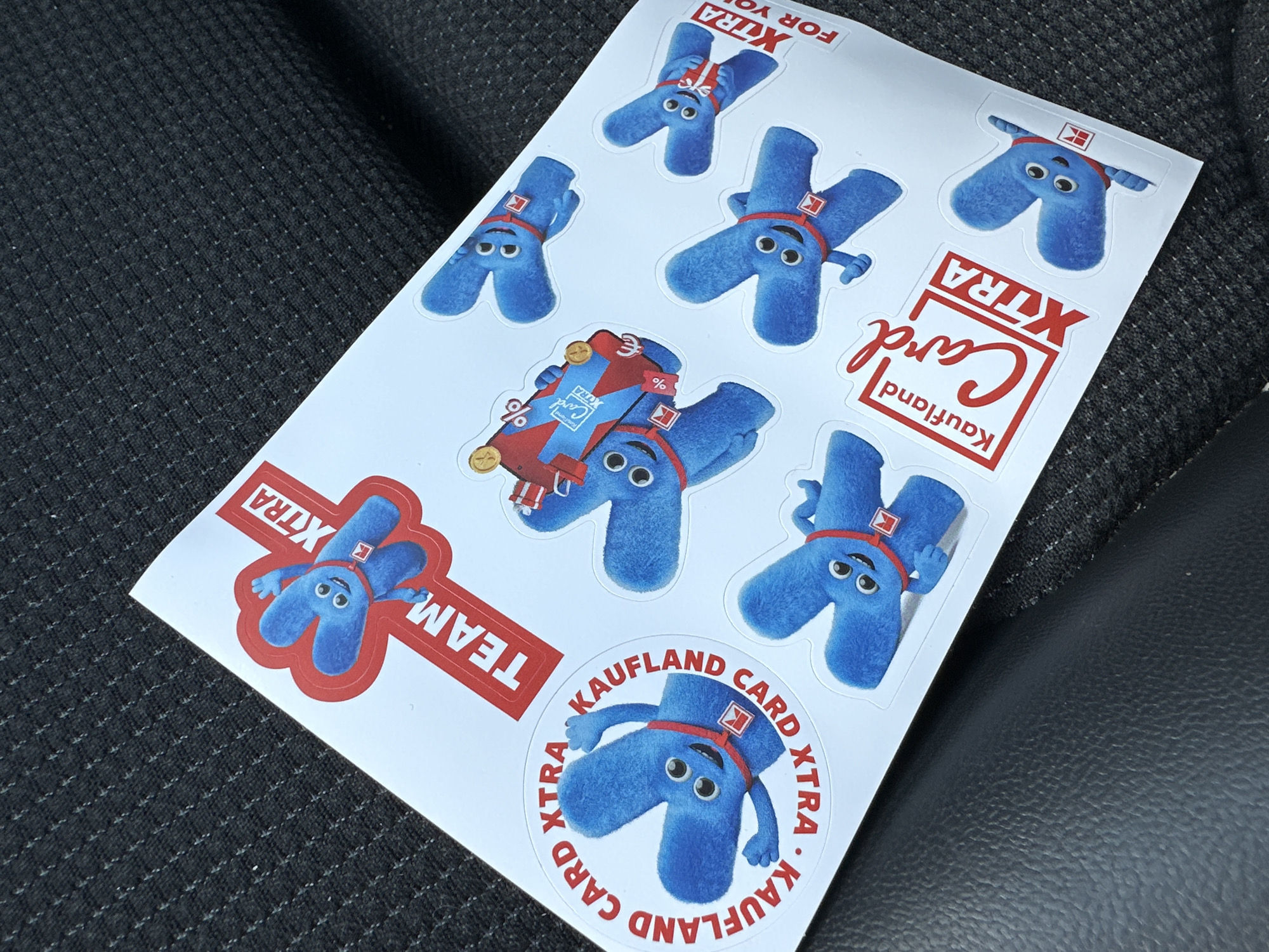 The image shows a sheet of stickers featuring a blue, furry character resembling the letter "X". Some stickers show the character holding a credit card, some show the character with a backpack, and some are within red shapes that display the words "TEAM" and "XTRA". Other stickers display the text "KAUFLAND CARD XTRA". The background appears to be a dark gray, textured material. It is not possible to determine if the location is Sofia, Bulgaria, as there are no elements in the image that provide any geographical context.