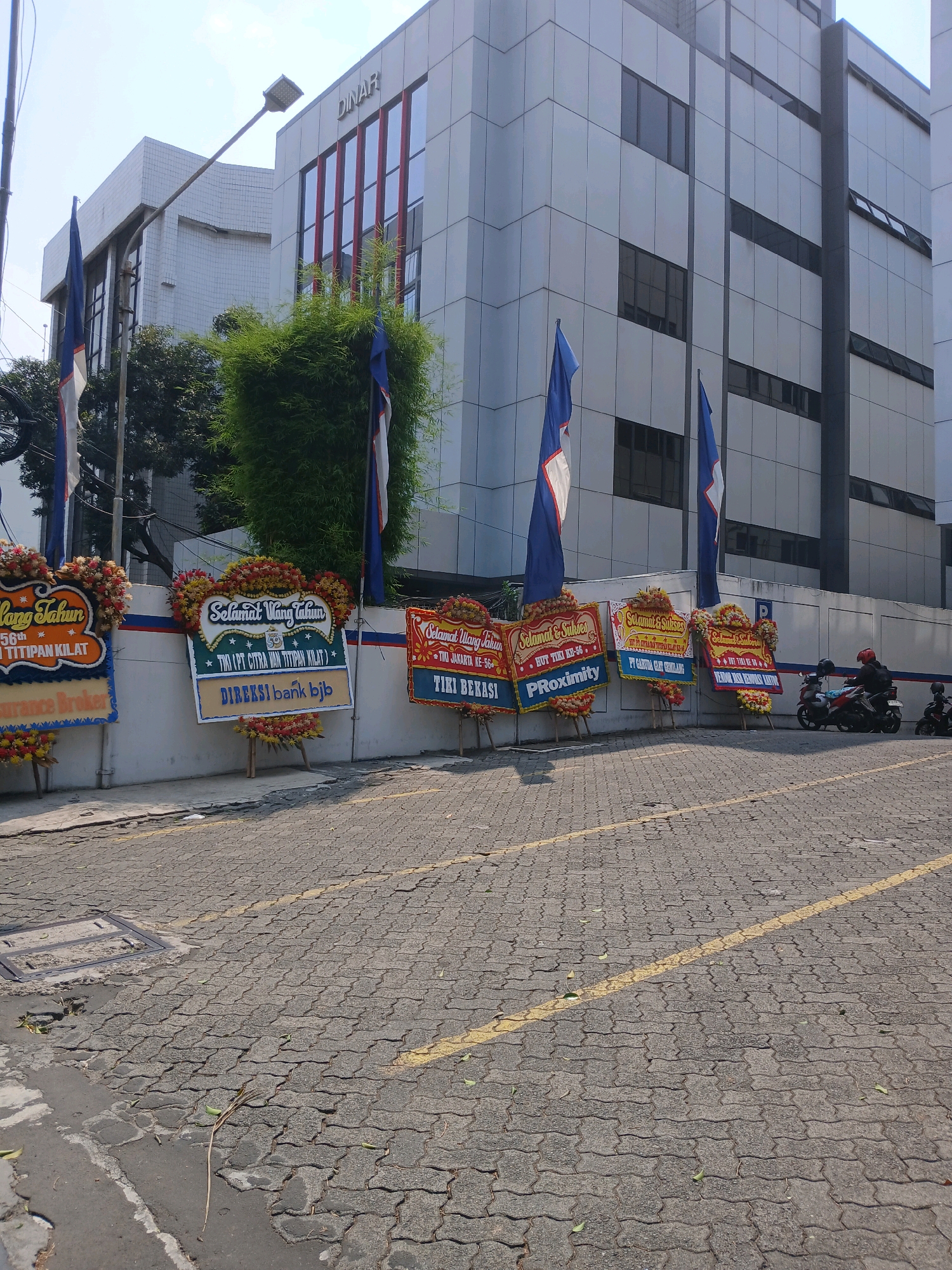 The image depicts the exterior of a modern, multi-story building with a gray facade and a prominent "DINAR" sign at the top. In front of the building, a white wall serves as a backdrop for several large floral arrangements, commonly known as "papan bunga" in Indonesia, which are typically used for congratulations or condolences. These arrangements are adorned with colorful flowers and contain various congratulatory messages, some referencing specific companies or events. Two blue and white flags are visible, attached to poles in front of the wall. The ground in front of the wall is paved with interlocking concrete blocks, with a yellow line marking a road or parking area. In the background, there are trees and other foliage. Further to the right, two motorcycles are parked, and one is being ridden by a person wearing a helmet. The scene suggests a public or commercial area in Central Jakarta, Indonesia, possibly in front of an office building or financial institution.