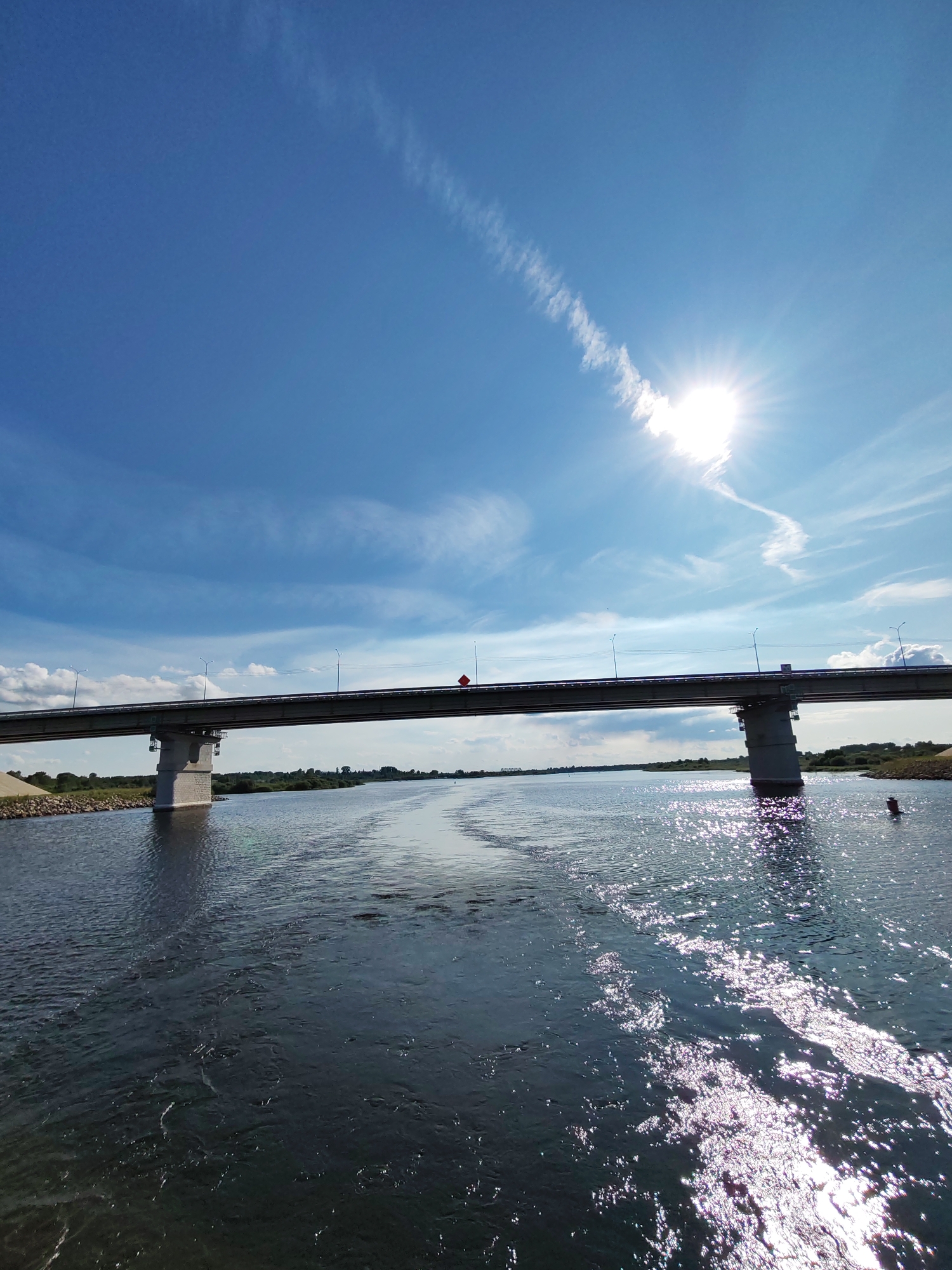 The image depicts a wide river with a bridge spanning across it. The sky is a bright blue with scattered white clouds and the sun is shining brightly, casting reflections on the water. The bridge has multiple lanes and is supported by concrete pillars. The riverbanks are lined with vegetation and some sandy areas. There are no people visible in the image, but there is a small red buoy in the water near the right bank. The overall scene suggests a sunny day with calm weather. This scene is located in the city of Nifantovo, Russia.