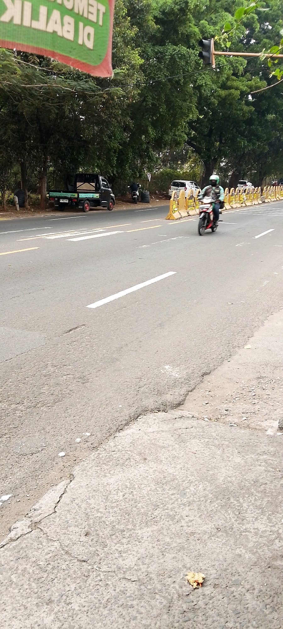 In Depok, Indonesia, a street scene unfolds. A motorbike with a rider wearing a helmet is moving down the asphalt road. The rider is positioned in the center-right of the frame. On the left side of the road, a black pickup truck is parked. Yellow traffic barriers are set up along the right side of the road, indicating a lane closure or construction. A traffic light is visible hanging above the road amidst the lush green trees that line the street. The road surface shows signs of wear and tear with cracks and a damaged edge visible in the foreground. A small piece of debris, possibly a leaf or food scrap, lies on the asphalt in the lower right corner of the image. A banner with text is partially visible in the upper left corner, adding to the urban context.