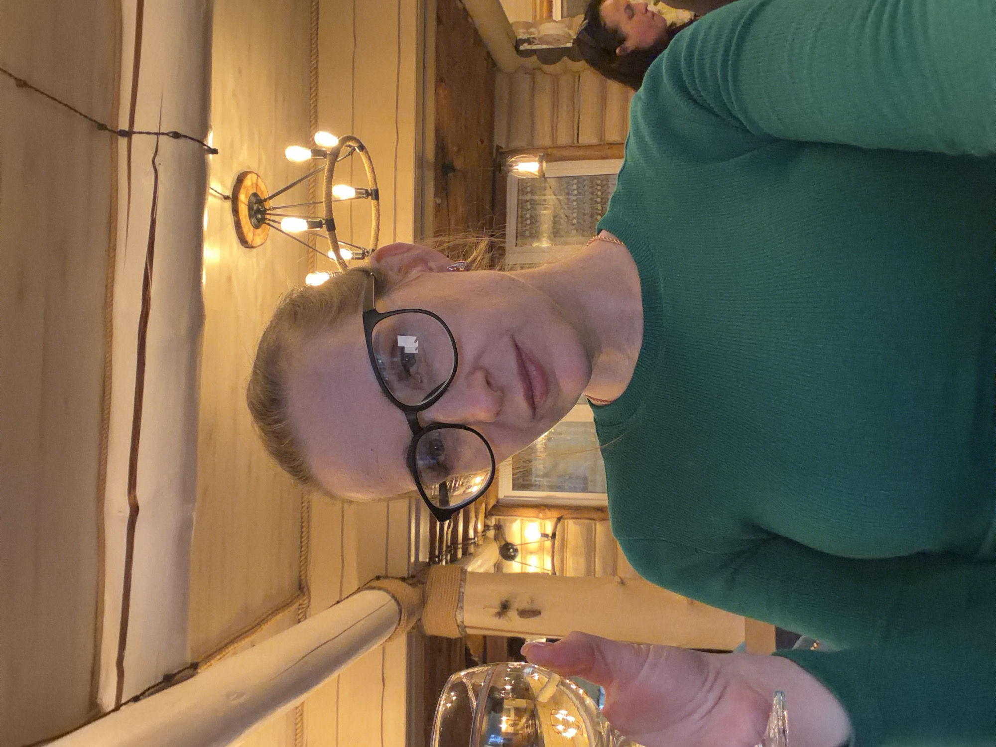 A woman wearing glasses and a green sweater is sitting at a table. She is holding a glass. The location is in the city of Zaplav'e, Russia. Behind her are wood walls, a chandelier, and a window. Another person is visible in the background.