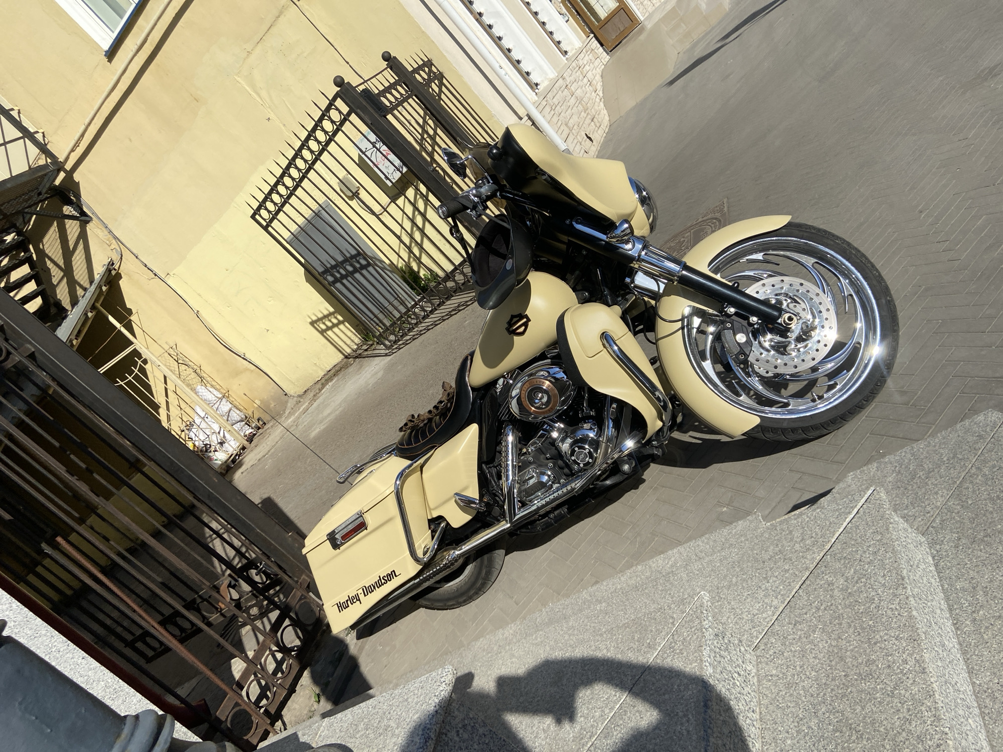 The image depicts a beige Harley-Davidson motorcycle parked on a paved surface, likely a sidewalk or alley, in an urban environment. The motorcycle is angled towards the viewer, showcasing its front wheel, engine, and fairing. The background includes a yellow building with a metal gate and a partial view of a white building. The lighting suggests it is daytime, with shadows cast on the ground. The scene is likely in Kazan, Russia, based on the architectural style and urban setting.