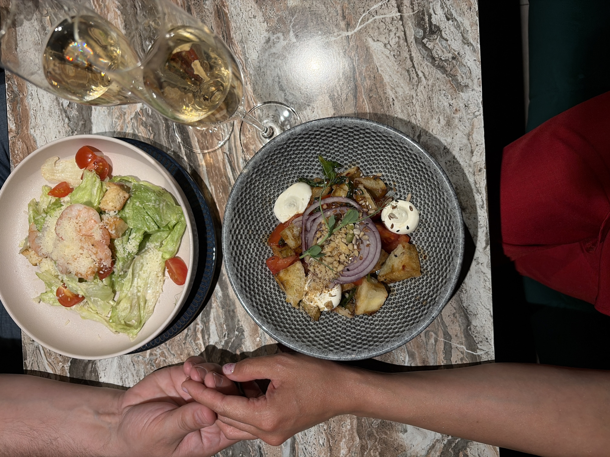 Overhead view of a dining table at a restaurant, possibly in Chaikovsky, Russia. Two champagne flutes filled with a light-colored sparkling wine sit on a table with a marbled surface. A light-colored bowl containing a salad with lettuce, tomatoes, croutons, and shredded cheese is placed to the left of the glasses. Beside it, a dark-colored bowl contains another salad with tomatoes, red onions, croutons, greens, and dollops of white sauce. Two people’s hands are clasped together near the bowls, suggesting a romantic or intimate dinner setting. A red fabric and a green surface appear on the right, likely from clothing or a seat.