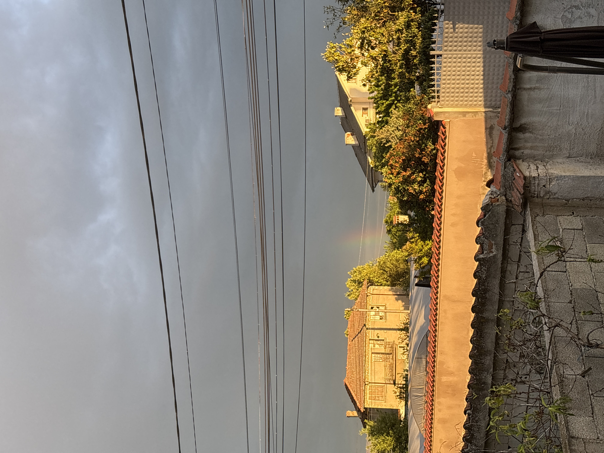 The photograph captures a view in Manole, Bulgaria. The sky is predominantly overcast with dark grey clouds. Electrical wires stretch across the upper left portion of the frame. In the distance, there is a faint rainbow visible. Buildings are on either side of the frame. The buildings are bathed in warm sunlight. One building is covered in greenery. A concrete wall is on the right, overgrown with a small tree.