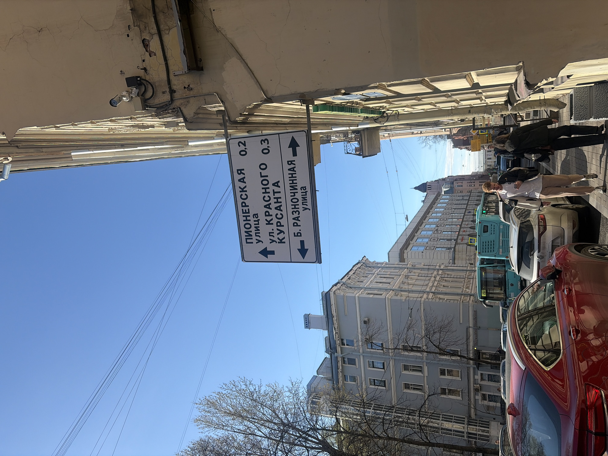 An eye-level shot captures a sunny urban street scene in Saint Petersburg, Russia, looking down a street lined with buildings and vehicles. The sky is a clear, bright blue, crisscrossed by multiple utility wires.
In the foreground, a prominent white street sign with black text is mounted to the underside of a building overhang. The text, in Cyrillic, indicates directions and distances: "ПИОНЕРСКАЯ улица 0,2" (Pionerskaya street 0.2 km) with an arrow pointing straight ahead; "УЛ. КРАСНОГО КУРСАНТА 0,3" (Krasnogo Kursanta street 0.3 km) with an arrow pointing left; and "Б. РАЗНОЧИННАЯ улица" (B. Raznochinnyaya street) with an arrow pointing right. Above the sign, the underside of the building shows weathered paint and some exposed wiring, with a security camera mounted.
To the right, a red car is prominently parked or moving slowly, partially obscuring a white sedan behind it. Further down the street, two blue and green city buses are visible in traffic. Several pedestrians are walking on the sidewalk to the right, including a young man in a white t-shirt and beige pants holding a phone or cup, and another person in a dark jacket.
The buildings lining the street are multi-story, featuring classical architecture with decorative elements and varying window styles. Some facades show signs of age, with cracked plaster and worn paint. Bare-branched trees are scattered along the sidewalk, suggesting either early spring or late autumn. The overall impression is a bustling daytime street with clear weather.