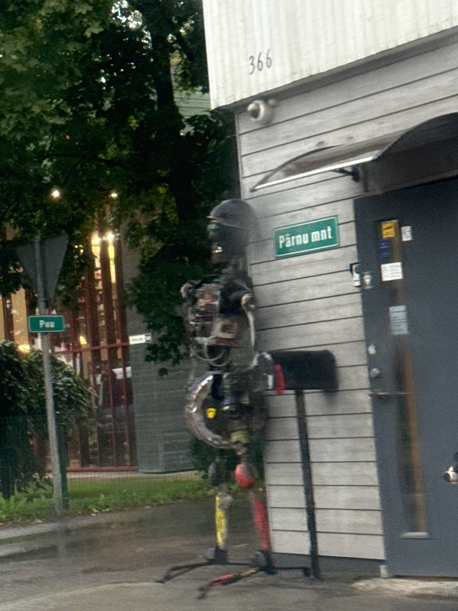 An outdoor, eye-level, medium shot shows a full-body, freestanding sculpture of a robot on a sidewalk in Tallinn, Estonia. The robot is constructed from various metallic parts, including a helmet, torso, and limbs. Its legs are painted yellow and red. To the left of the robot, a street sign with green lettering reading "Puu" is visible. Behind the robot, a dark gray door with several stickers and a mailbox with a red flag is attached to a building with horizontal gray siding. The building has the number "366" painted in black above the door. A security camera is mounted on the wall. The background includes lush green trees and a chain-link fence. The ground is wet, suggesting recent rain. The overall lighting is dim, with some dappled sunlight filtering through the trees.