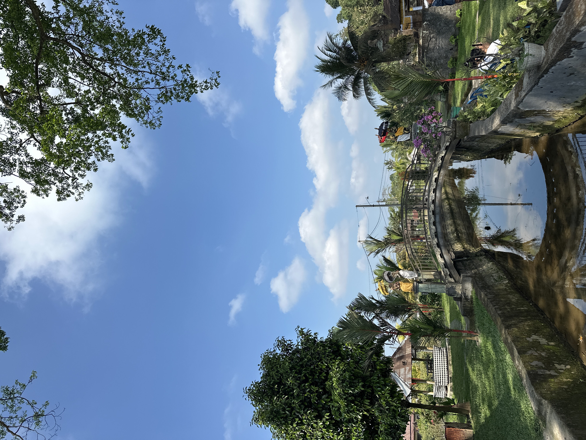The image depicts a scenic outdoor setting, likely in Marga, Indonesia, under a bright blue sky dotted with scattered white clouds. A gently arched bridge, constructed of what appears to be stone and possibly wood, spans a narrow, dark-colored canal filled with calm water that reflects the sky and surrounding elements. The canal banks are lined with weathered concrete walls, partially covered in moss.
To the left of the canal, a lush green lawn is bordered by trees. One prominent tree with dense foliage stands tall, while other smaller palm-like plants are visible. A rustic gazebo with a corrugated metal roof, featuring a checkered pattern draped over its seating area, is also present on the left.
On the bridge, two statues stand on stone pedestals. The statue on the left wears a yellow garment, and the one on the right wears a white and yellow garment. The bridge is decorated with railings and a cluster of vibrant purple flowers in a pot on the right side.
To the right of the canal, more greenery is present, including palm trees and potted plants. In the background, glimpses of buildings with traditional architecture, possibly temples or residences, can be seen. Several people are visible in the background on the right, some appearing to be seated or standing near the canal. A power line with a utility pole stretches across the upper right portion of the scene. The overall impression is one of a tranquil, possibly culturally significant, outdoor space.