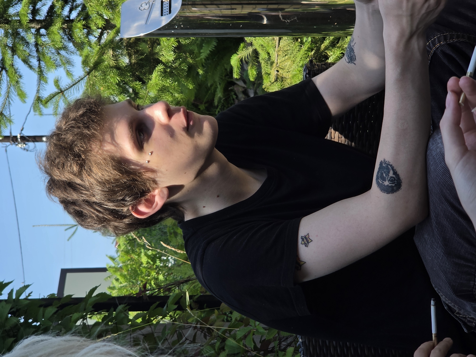 A medium shot captures a young person with light brown, tousled hair, wearing a black t-shirt, sitting outdoors. They have two silver studs on their left cheek. Their left arm is resting on their lap, revealing a tattoo of a star with radiating lines on their upper arm and another tattoo of a circular, shadowy design on their forearm. The person is holding a lit cigarette in their right hand, with their fingers and thumb positioned around the filter and body of the cigarette. The background consists of lush green foliage and trees, with a clear blue sky overhead. To the right, a silver cigarette receptacle is visible, featuring a prominent cigarette logo. The scene suggests a relaxed, outdoor setting, possibly a park or a patio, given the presence of the receptacle. The location is identified as the city of Nagorny, Russia.