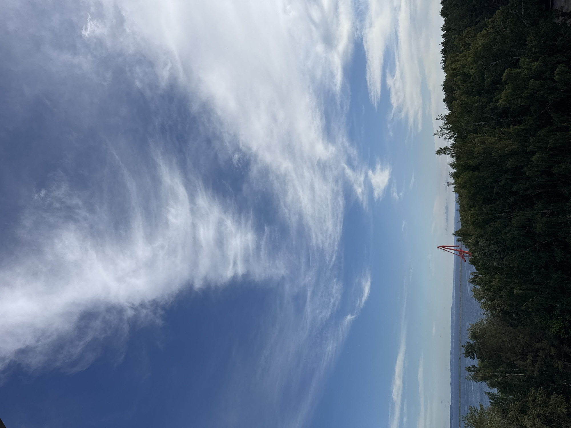 A wide-angle, high-angle shot captures a scenic view of the Kama River in Chaikovsky, Russia, under a dynamic sky. The upper half of the frame is dominated by a brilliant blue sky with wispy cirrus clouds streaking across it in a graceful, undulating pattern. Below the sky, a dense, dark green forest forms a thick canopy, stretching across the lower portion of the image. The trees appear lush and varied, with some lighter-colored foliage interspersed amongst the darker shades.
The Kama River, a wide expanse of calm, blue water, occupies the middle ground, reflecting the sky above. Along the distant horizon, a low, hazy line suggests rolling hills or the opposite shore of the river. Prominently positioned on the right side of the riverbank, emerging from the treeline, is a tall, red industrial crane. Its angular structure stands out against the natural landscape, hinting at port or construction activity. The scene is tranquil and bathed in natural light, suggesting a clear or partly cloudy day. There are no people visible in the image.