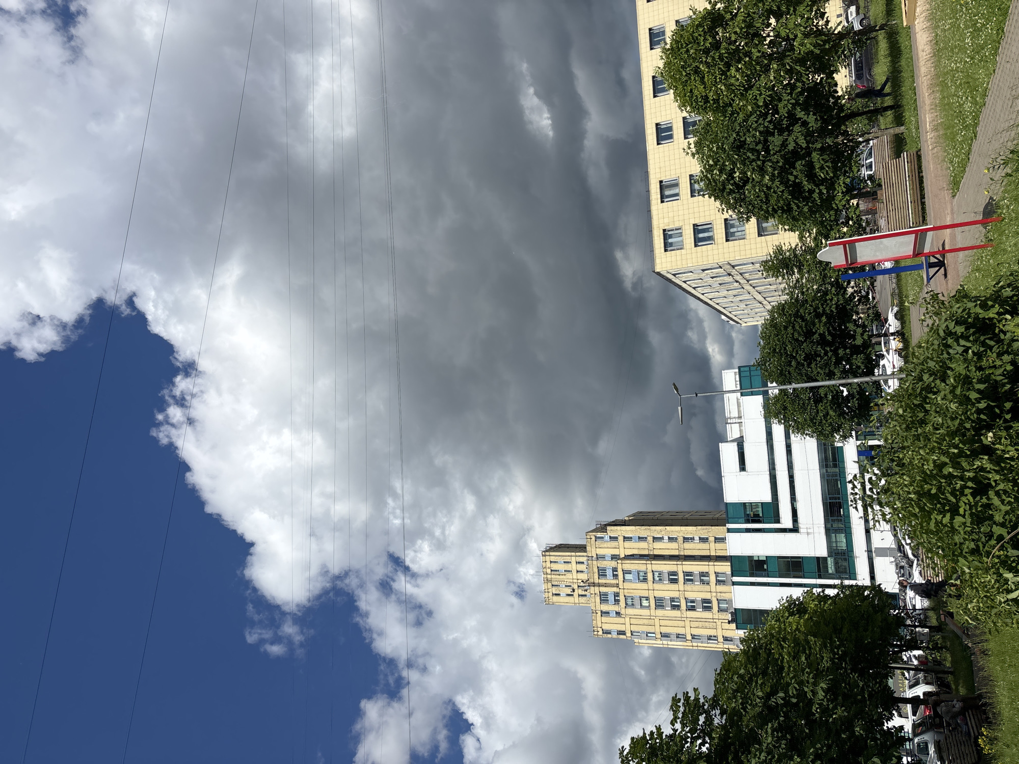 This media file captures a street-level view of urban buildings against a dramatic sky. The scene is oriented vertically, suggesting it was filmed or photographed in portrait mode.
Dominating the upper portion of the frame is a sky filled with a mix of dark, ominous storm clouds and bright, fluffy white clouds, with patches of clear blue sky visible. A series of parallel overhead wires traverse the sky, adding a linear element to the composition.
Two distinct buildings are visible. The building on the right is a multi-story structure with a pale yellow facade, punctuated by numerous windows. Trees with lush green foliage partially obscure this building. To the left and slightly further back, another building with a white and green facade is visible, appearing more modern in design.
In the foreground and to the right, there are elements of an outdoor space. A paved walkway with grass and small white flowers lines the edge. A wooden bench is present, and beyond it, several cars are parked along the street. The presence of a streetlamp indicates it is an outdoor urban environment.
The weather appears changeable, with the dark clouds suggesting an approaching storm or recent rainfall, contrasting with the bright white clouds. The time of day is likely daytime, given the natural light illuminating the scene. The visible text on a partial sign in the foreground reads "ДАРЕН". The overall mood is somewhat somber due to the stormy sky, yet the presence of trees and a bench suggests a public or residential area. The overall setting appears to be a street in Saint Petersburg, Russia.