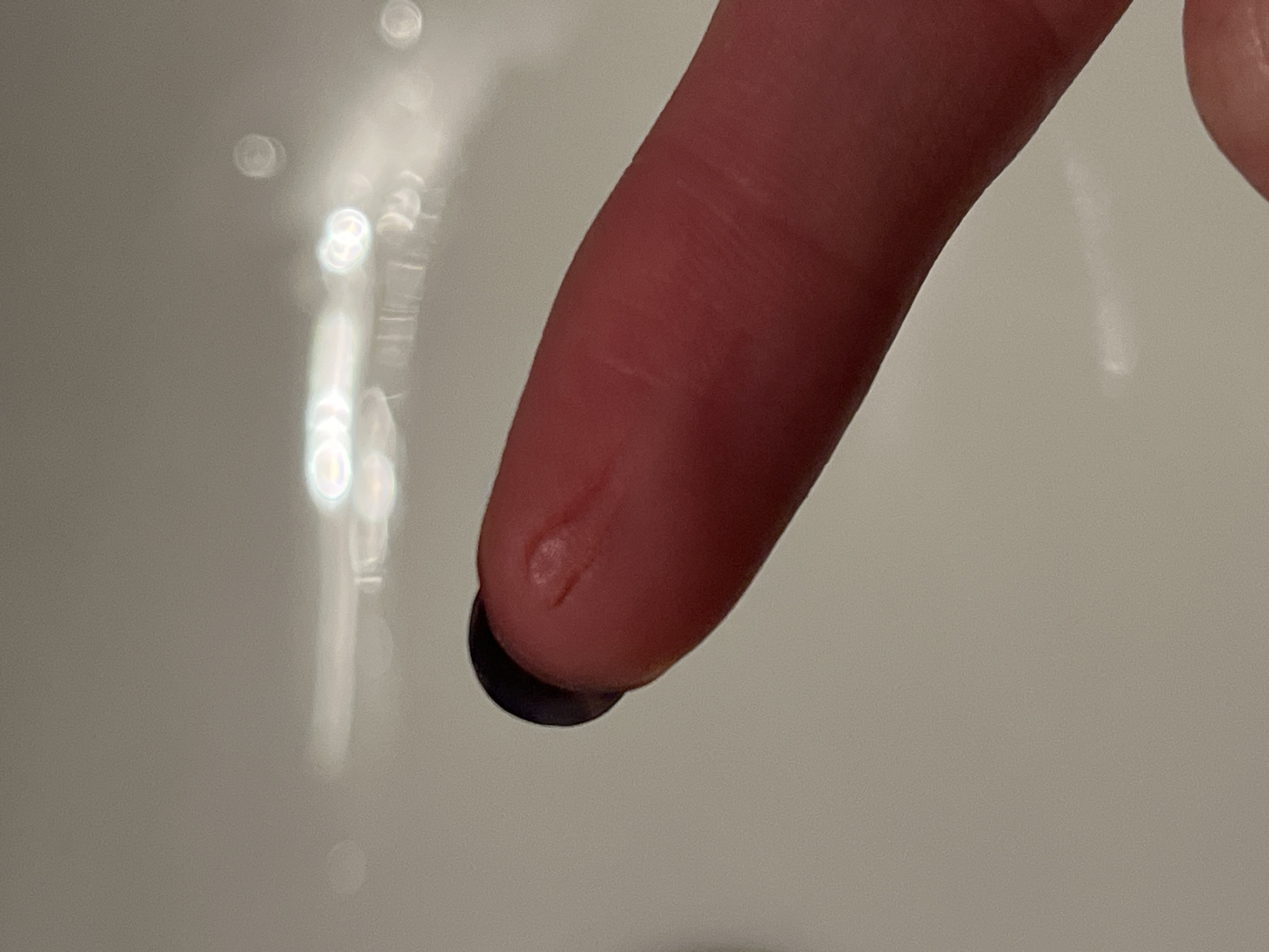 A close-up, high-angle shot focuses on a human fingertip holding a small, dark, circular object. The finger, likely an index finger, is oriented diagonally across the frame, with its tip facing downwards towards the bottom left. The skin of the finger appears light to medium tone, and there are some minor marks or imperfections visible on the pad of the fingertip.
The small object held by the finger is dark, almost black, and appears flat or slightly convex, resembling a small bead, seed, or a tiny electronic component.
The background is a light, monochromatic surface, possibly a sink, bathtub, or another ceramic or reflective surface. Out-of-focus, blurry circular and elongated bright reflections are scattered across the background, suggesting the presence of overhead lighting or reflections from a wet surface. These reflections create a soft bokeh effect.
The scene appears to be indoors, likely in a well-lit room, but the exact location (such as San Bernardino Tlaxcalancingo, Mexico, as provided) is not visually discernible from the image. No specific activity beyond holding the object is clear, nor are any emotions or visible text present.