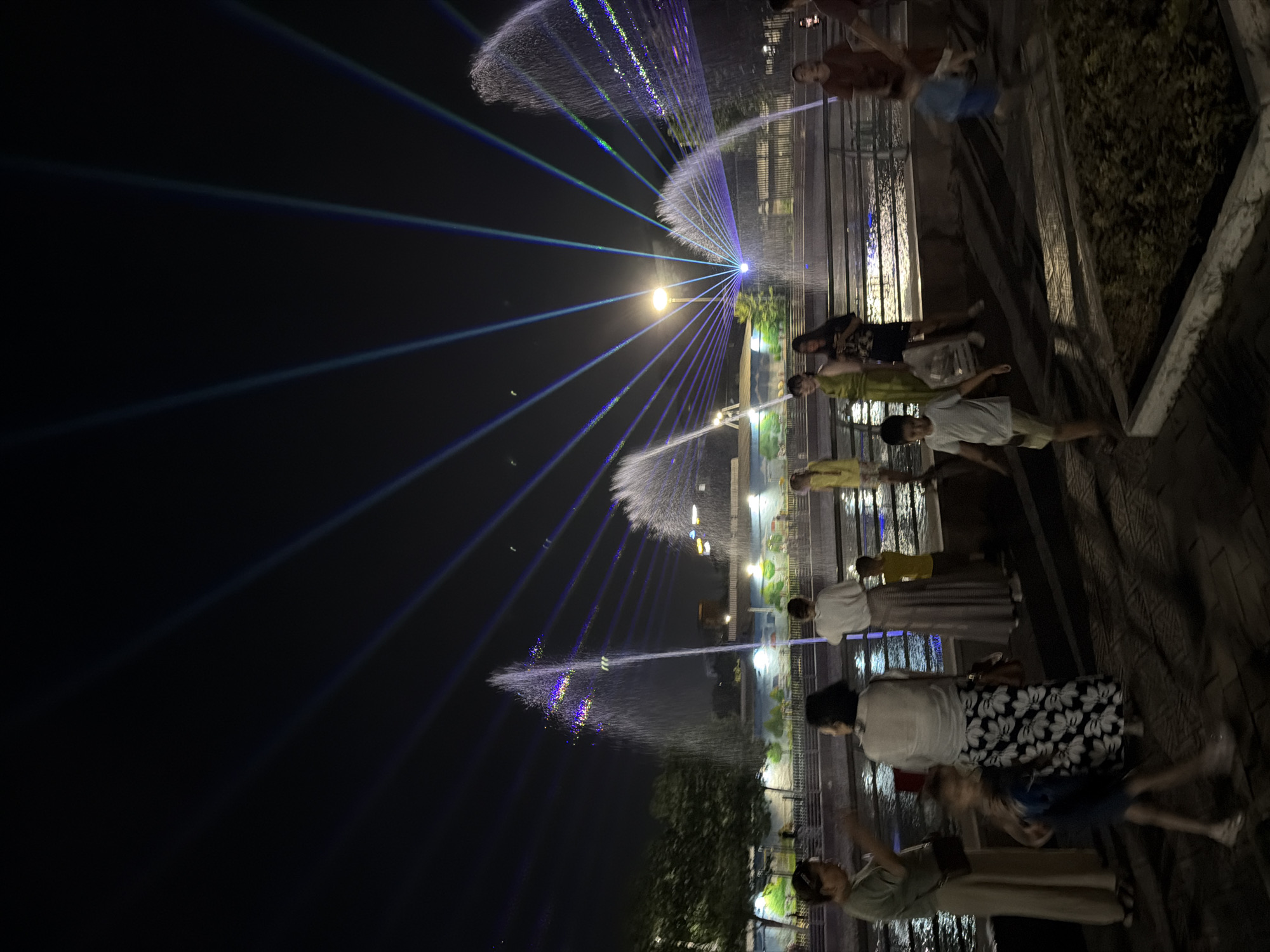 The image depicts a night scene with a water fountain show and laser lights. Several people are gathered, standing and watching the spectacle. The fountain jets are illuminated with blue and green lights, creating a dramatic effect against the dark sky. Laser beams radiate from a central point, crisscrossing through the water sprays and forming intricate patterns. Some of the people appear to be families, with adults and children present. The setting suggests an outdoor entertainment area, possibly a park or plaza, with a body of water where the fountain is located. The time of day is clearly night, as indicated by the dark sky and artificial lighting. The weather appears clear, allowing for the light show to be fully appreciated. There is no visible text in the image. The overall mood is one of enjoyment and wonder, as people are captivated by the visual display. The orientation of the image is vertical, with the fountain show dominating the left side and the viewers on the right. The location context of Shymkent, Kazakhstan, does not provide specific visual cues within the image itself.