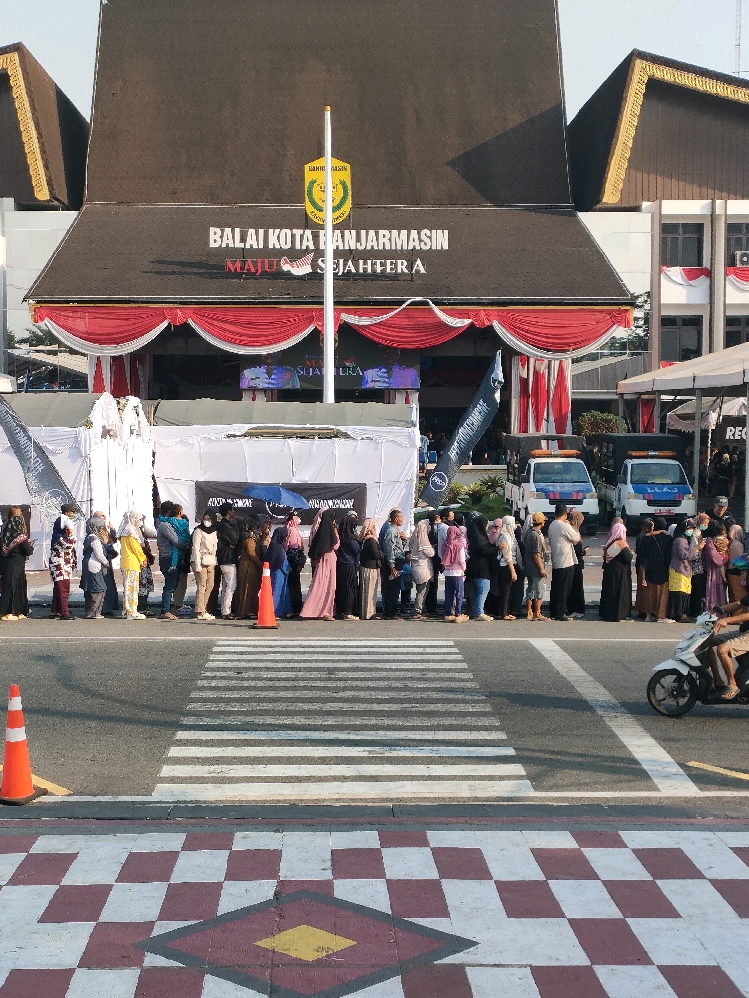 The image depicts the exterior of the Banjarmsin City Hall building in Banjarmasin, Indonesia, during the daytime. A large crowd of people, many wearing hijabs and modest clothing, are lined up along the sidewalk in front of the building, suggesting an event or a queue for services. The building's facade is decorated with red and white drapes, and a large banner with Indonesian text is visible. Two white utility vehicles with blue stripes are parked behind the crowd, and a security guard is visible near one of them. A white motorcycle with a rider is in the foreground, crossing a pedestrian crosswalk. The ground features a checkered pattern of red and white tiles leading to the crosswalk. The sky is clear and bright. The overall scene suggests a public gathering or official function at the city hall.