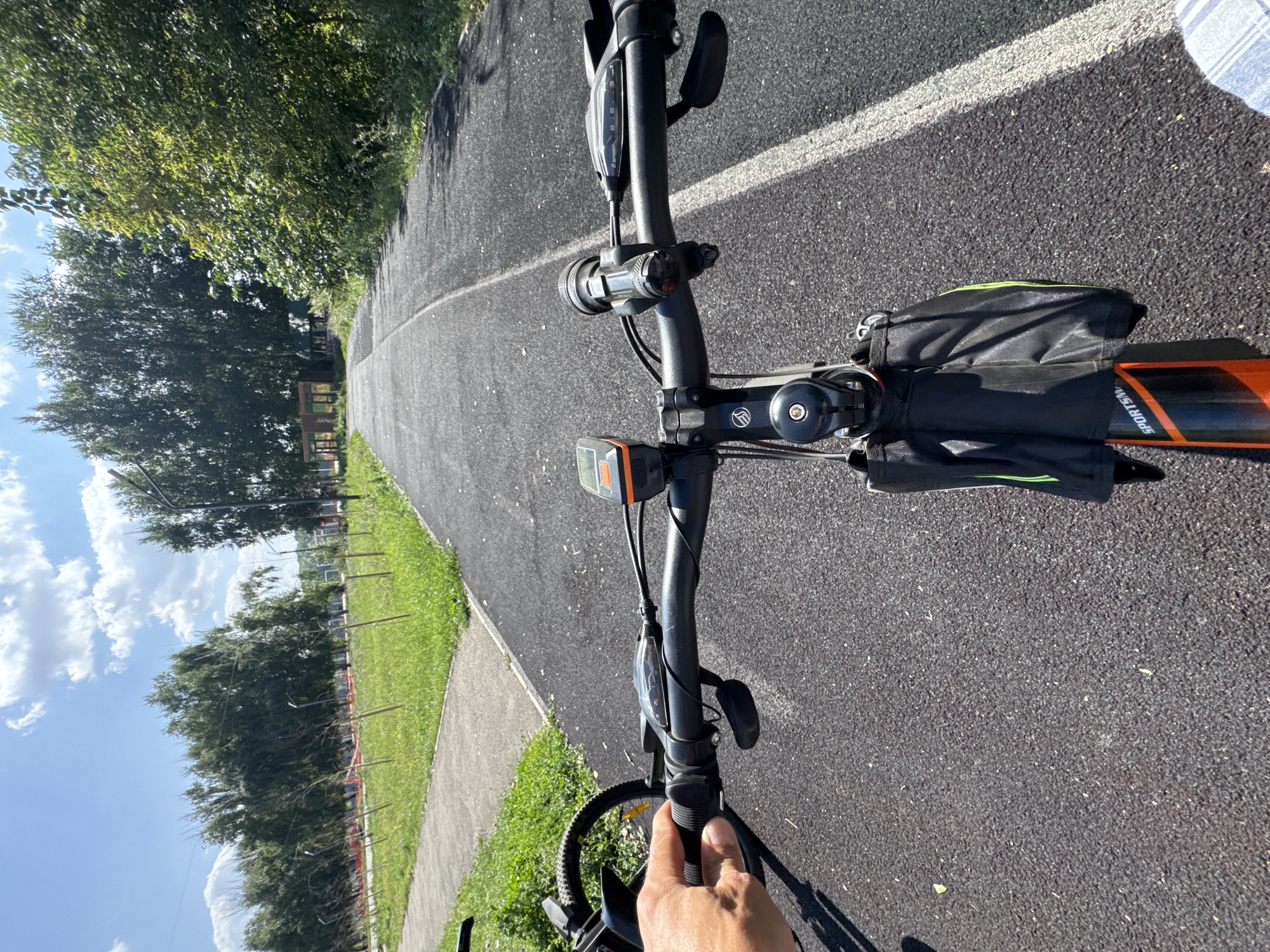 The image shows a first-person perspective of someone riding a bicycle on a paved path, likely a bike trail or sidewalk, in a park-like setting. The path is dark asphalt with a white line marking its edge on the right. To the left of the path, there is a strip of green grass with some trees and saplings, many of which are supported by wooden stakes. Further to the left, there's a paved walkway. The background features a dense line of trees and a clear blue sky with scattered white clouds. In the distance, a small wooden structure with windows is visible amongst the trees. The bicycle's handlebars are prominent, with a hand gripping the right handlebar. Attached to the handlebars are a bike computer and a headlight. A small saddlebag is mounted on the bike's frame below the handlebars. The overall scene depicts a sunny day, suitable for outdoor recreational activities. The location is identified as the city of Oskemen, Kazakhstan.