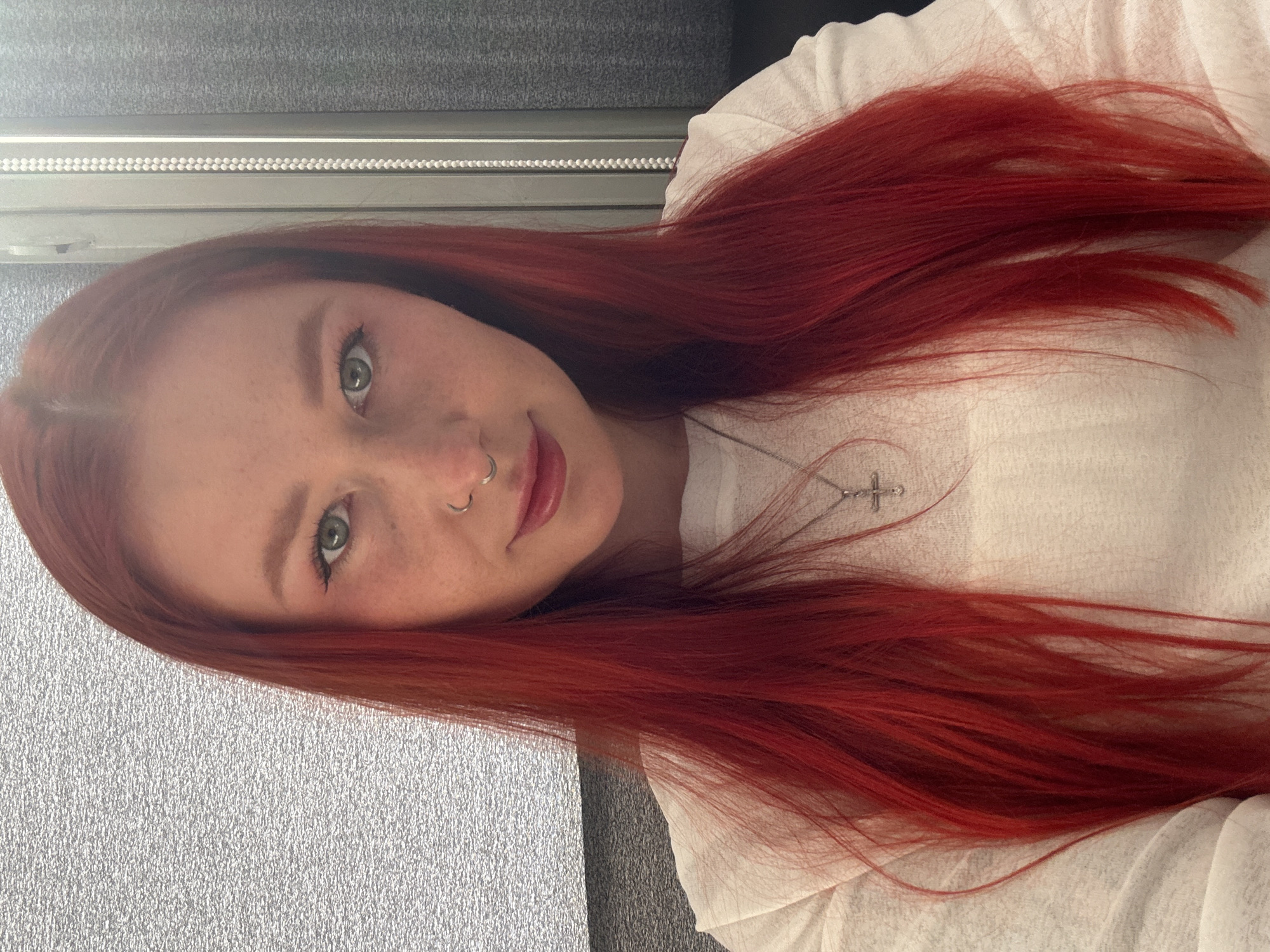 A young woman with striking, vibrant red hair is captured in a close-up, slightly tilted selfie. She looks directly at the camera with a soft, neutral expression and a hint of a smile. Her eyes are a distinct green, and her fair skin shows subtle freckles across her nose and cheeks. She has a silver septum piercing in her nose.
She is wearing a simple white top, and a delicate silver necklace with a small cross pendant is visible against her chest, partially obscured by her long hair. Her voluminous, bright red hair frames her face and extends downwards, showcasing its rich color and texture.
The background is simple and indoors, featuring a grey, textured wall or surface on the left side of the frame. Behind her head, there's a horizontal band of a darker grey material, possibly a window blind or part of a furniture piece, decorated with a row of small, light-colored dots. The lighting is soft and even, suggesting an indoor setting without harsh shadows.
The overall impression is a casual portrait or selfie, focusing on the individual's features. There are no visible location cues within the image to suggest its context in Rostov-on-Don, Russia, nor is any text visible.