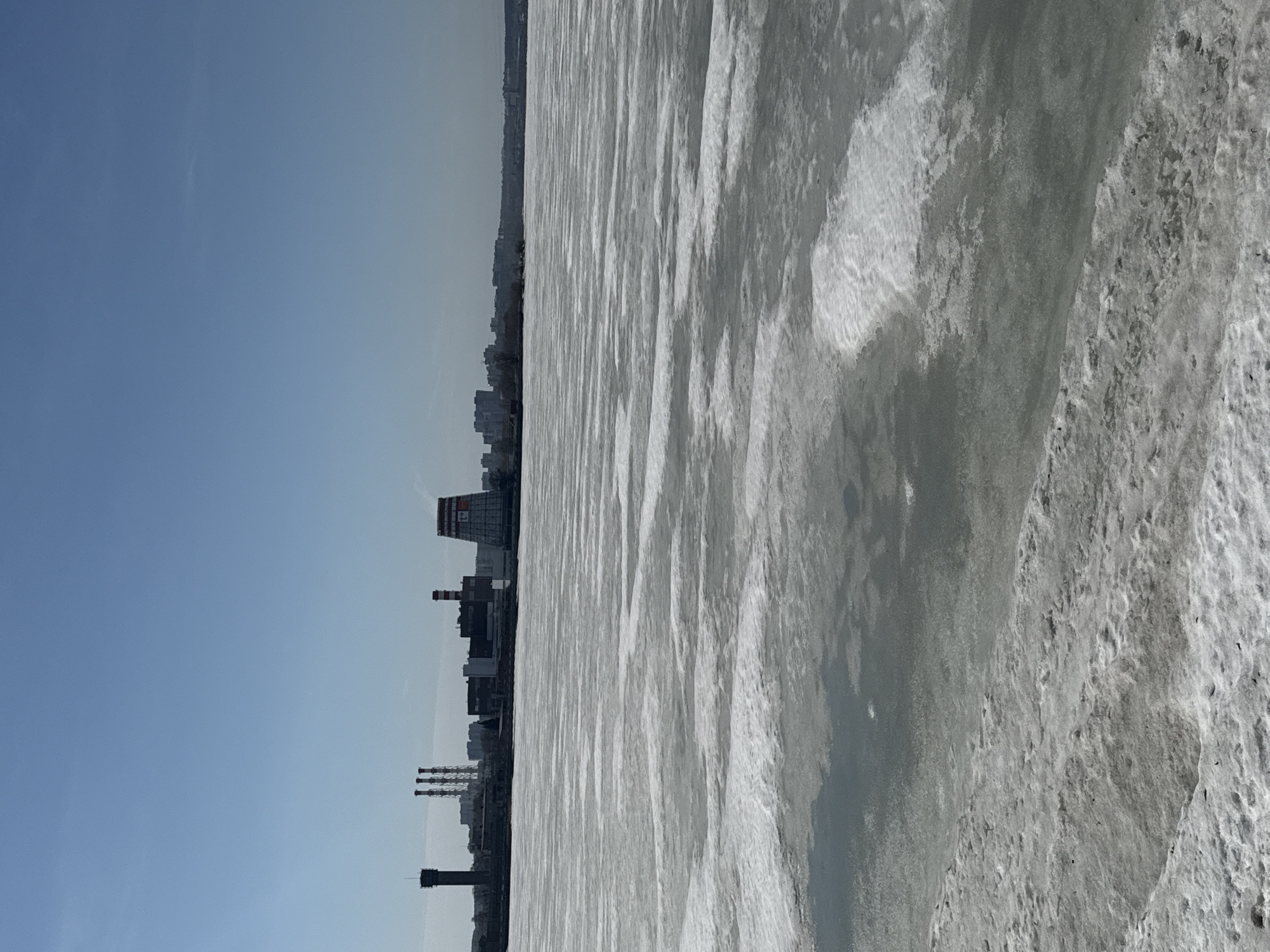 The image captures a frozen section of the Izhevsk reservoir in Russia, with the city skyline visible in the background. The foreground is dominated by the textured surface of the frozen water, exhibiting shades of gray and white, indicating ice and snow cover. The city skyline is characterized by industrial buildings and smokestacks, along with residential buildings. The sky is a clear, light blue.