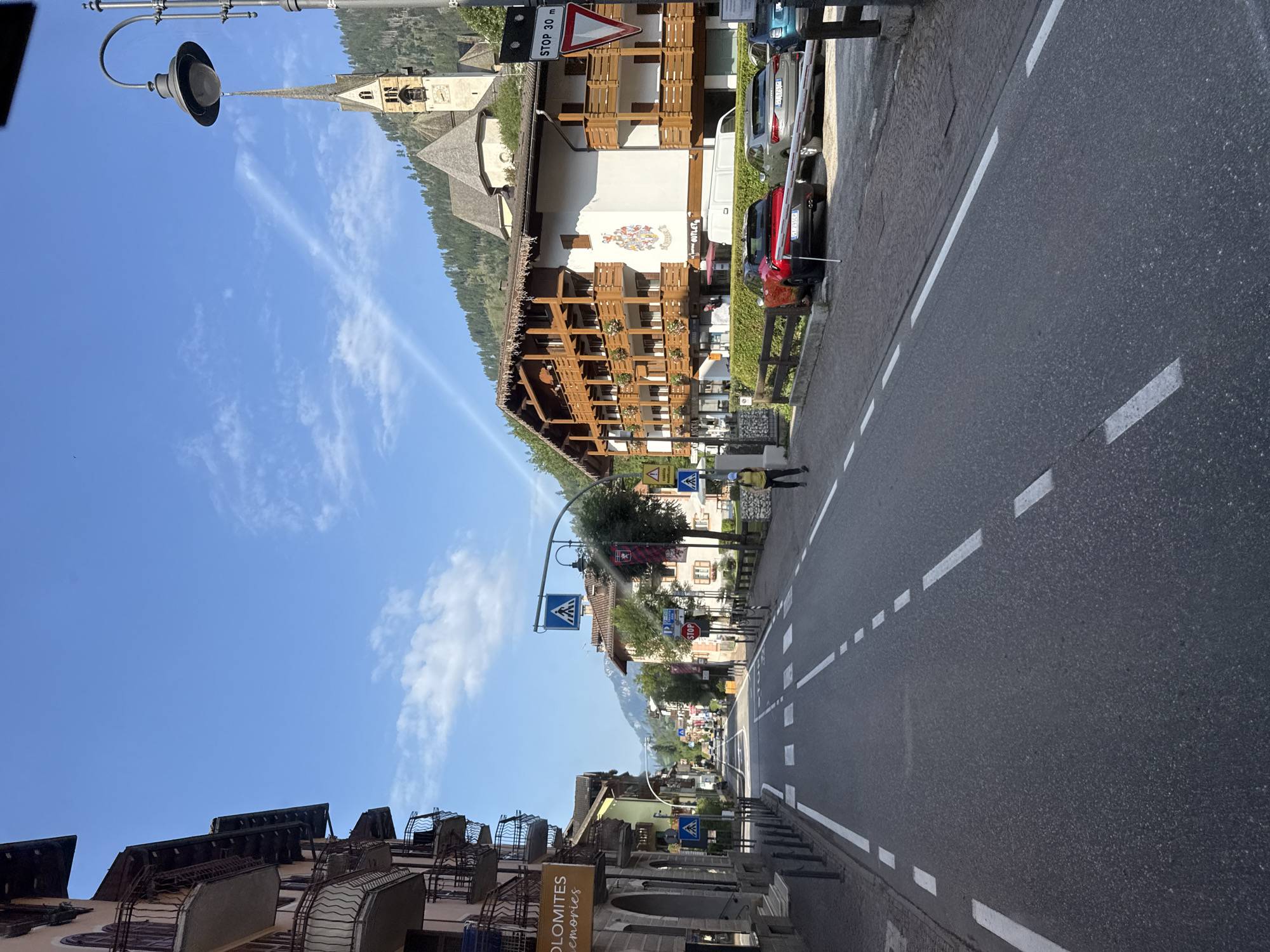 The image depicts a street scene in Moena, Italy, on a clear, sunny day. A two-lane asphalt road runs through the center of the frame, marked with white lines indicating lanes and pedestrian crossings. On either side of the road are multi-story buildings with traditional alpine architecture, featuring wooden balconies and pitched roofs.
To the left, a building displays a sign that reads "DOLOMITES memories." Further down the street, on the right, a prominent church with a tall spire is visible, set against a backdrop of forested hills. Streetlights and various traffic signs, including a "STOP 30 m" sign, are present along the road.
Several cars are parked along the right side of the street, and a person wearing a yellow top and dark pants is walking on the sidewalk on the right. The overall impression is of a peaceful, well-maintained town center.