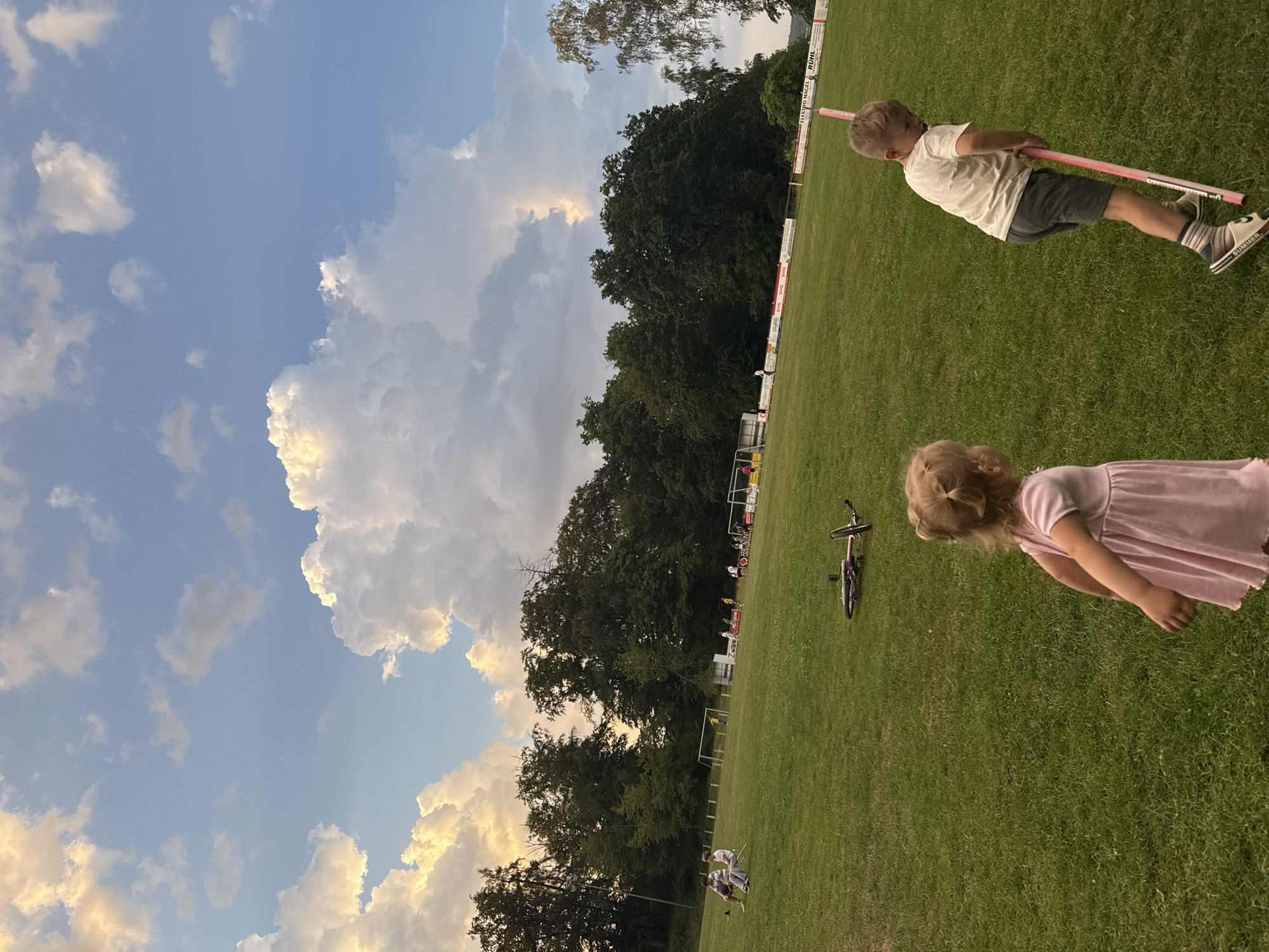 In Lumda, Germany, this image captures a grassy field with a treeline in the background. In the foreground, a young boy in a white t-shirt and grey shorts stands holding a pink pole, and a young girl in a pink dress stands to his left, facing away from the camera. Further in the background, several people are visible, some sitting and others standing near soccer goals. A bicycle lies on its side in the middle of the field. The sky above is partly cloudy, with dramatic, sunlit cumulus clouds.