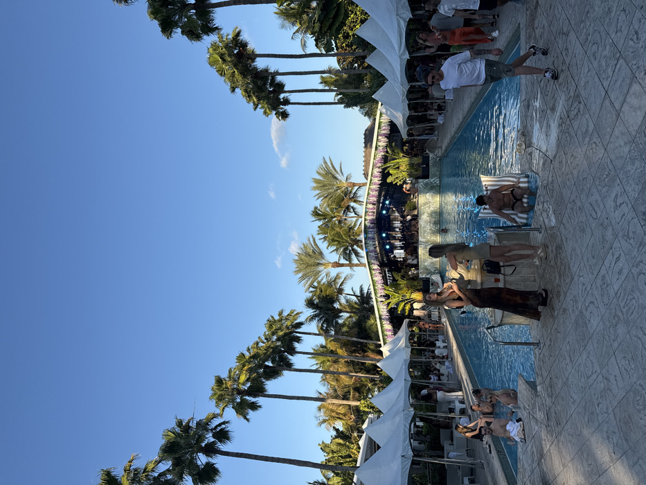 This image depicts an outdoor pool area during a late afternoon or early evening event, under a clear blue sky, located at a resort or hotel in Miami Beach, United States of America, consistent with venues such as The Fontainebleau Miami Beach.
The scene is dominated by a large, rectangular swimming pool with light blue water and white coping, extending from the midground towards the background. The foreground consists of a light-colored, patterned tiled patio. To the left of the pool, a line of six white, peaked canopy tents provides shaded seating areas, with several individuals seated beneath them on white lounge furniture.
In the background, a prominently featured stage structure with a large canopy roof stands behind the pool. The stage's backdrop is a light blue tiled wall, and its canopy is extensively decorated with hanging purple floral elements, suggesting wisteria. The stage is illuminated and appears to have people on or around it. The entire periphery of the scene and the distant background are densely populated with numerous tall palm trees of various species, characteristic of a tropical environment.
Approximately 30 to 40 individuals are visible, primarily adults, engaged in various social activities. In the immediate midground by the pool's edge, two women stand near a light-colored decorative fixture; one wears a long, dark patterned dress and holds a glass, while the other wears a light-colored two-piece outfit. To their right, a man sits upright on a white and gray striped inflatable lounge chair in the pool. Other people are observed standing in the shallow end of the pool, sitting on its edges, or mingling on the surrounding patio. On the right side of the patio, a man wearing a white t-shirt, patterned shorts, sneakers, and a blue bandana walks from right to left. People are dressed in casual resort wear, swimwear, and light clothing. Long shadows are cast from the left side of the image onto the tiled patio, consistent with low-angle sun.