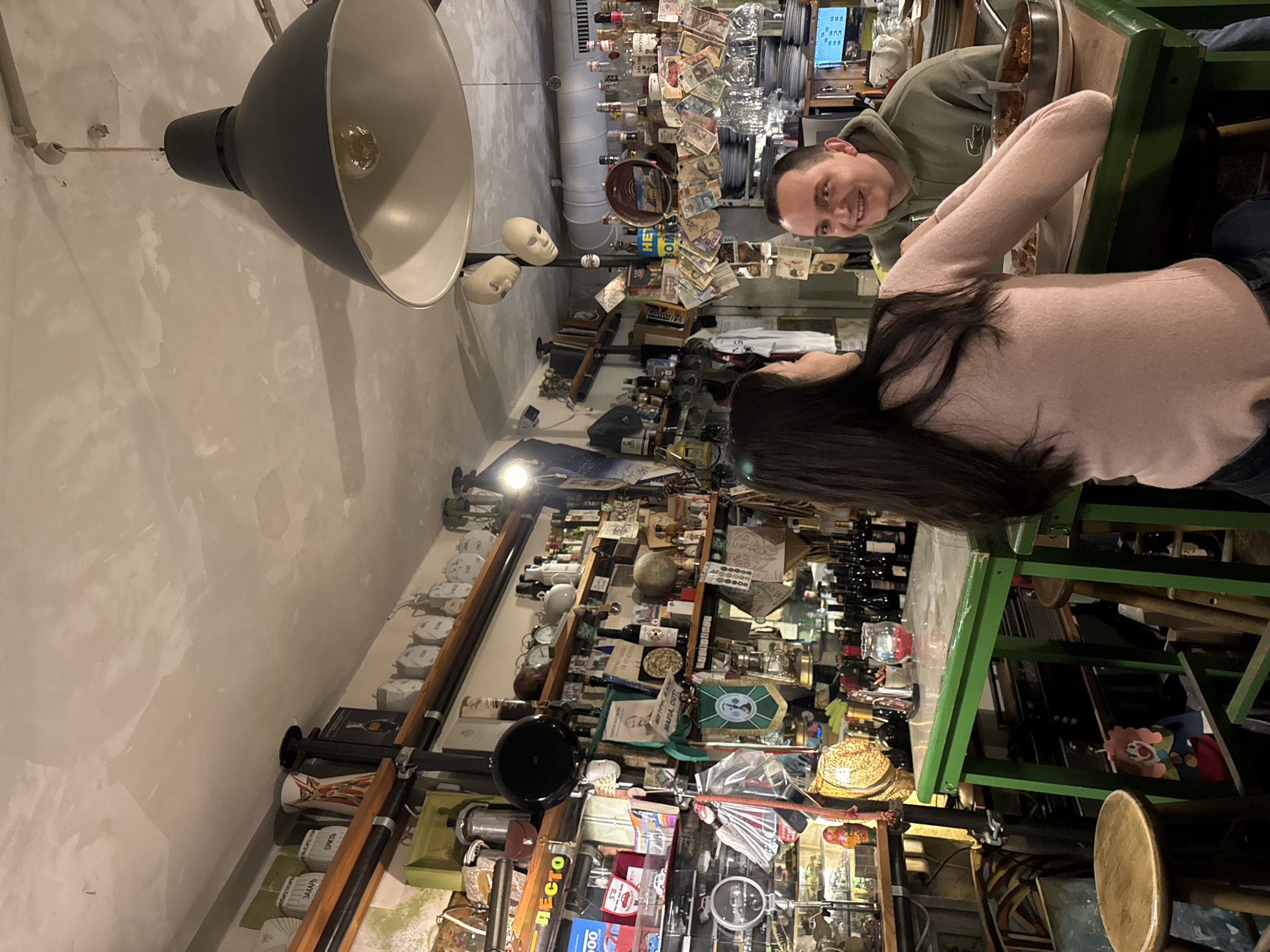 The scene depicts the interior of a highly cluttered and eclectic bar or restaurant located in Sofia, Bulgaria. Two individuals, an adult male and an adult female, are seated at a tall, green-framed table with a light-colored surface. The female, with long dark hair, is positioned with her back partially to the camera, wearing a light pink long-sleeved top and dark pants, her forearms resting on the table. She is turned towards the male. The male, positioned opposite her, has short dark hair and wears a green hooded sweatshirt featuring a visible Lacoste logo; he is smiling and looking directly at the female.
On the table between them, a metal pan containing a dark stew-like dish is in front of the male, and a white plate is partially visible near the female. A small red candle holder with a lit candle, a silver shaker, and a small glass jar are also present on the table surface. Wooden stools are visible beneath the table.
The surrounding walls and shelving units are densely packed with a vast array of miscellaneous objects, creating a maximalist aesthetic. These include numerous white ceramic storage jars, some with Cyrillic labels such as "САХАР" (sugar) and "ОРИЗ" (rice), decorative plates, various bottles of alcohol (wine and spirits), small model vehicles, vintage items like clocks and scales, a black pan, a metal coffee pot, framed pictures (one reading "Primo Libro di Mafia, Out!"), decorative globes, green banner-like insignias, and a multitude of foreign banknotes taped to a vertical surface near the bar area. Two featureless white masks hang from the ceiling near the bar.
The ceiling is light-colored and unevenly textured. Lighting is provided by a large, dark grey industrial-style pendant lamp with a white interior, hanging prominently in the upper right foreground, and another exposed bulb fixture on the left. The background features a bar area with stacked white plates, hanging glassware, and numerous liquor bottles. A ventilation duct is visible above the bar. A clown-themed decoration hangs beneath the table on the left. The overall lighting is warm and ambient.