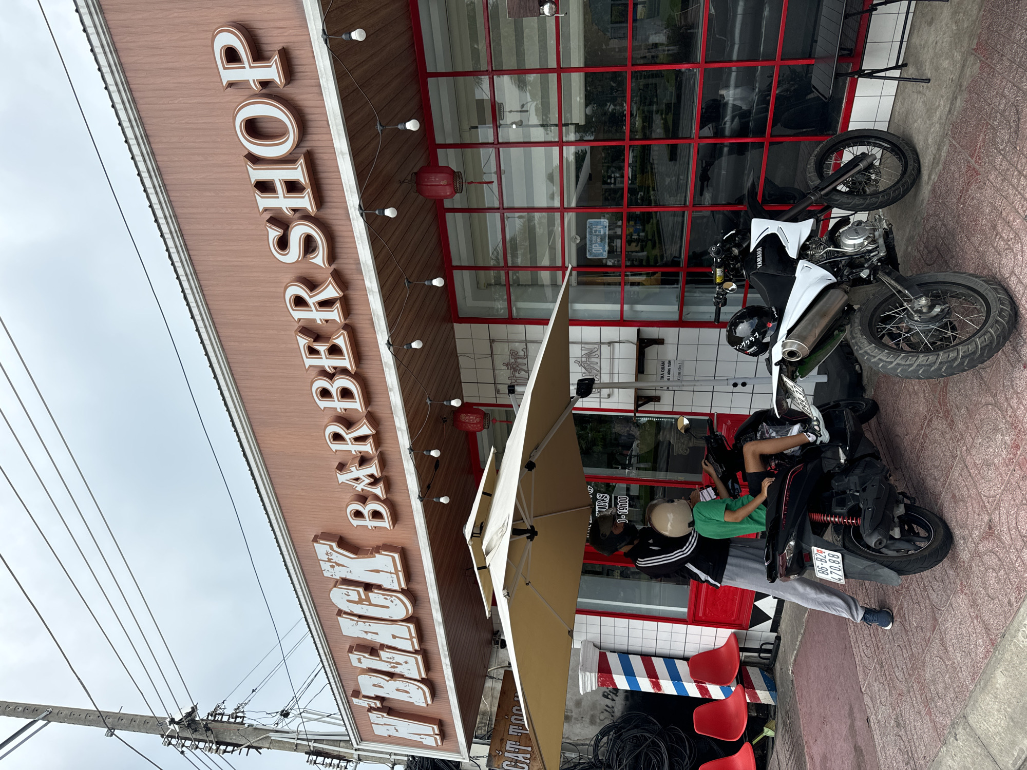 The image depicts the exterior of "N'BLACK BARBER SHOP" in Phan Thiết, Vietnam. The shop has a prominent sign with raised white lettering against a wood-grained facade. The building features large windows with red frames. In front of the shop, a beige umbrella shades a scene where an adult and a child are on a black scooter. The adult, wearing a black jacket with white stripes and grey pants, stands next to the scooter, while the child, in a green shirt and helmet, is seated on it. Another white motorcycle is parked to the right of the scooter. Red chairs are visible to the left, and a traditional barber pole stands near the entrance. Overhead wires and a utility pole are present in the background. The pavement is tiled.