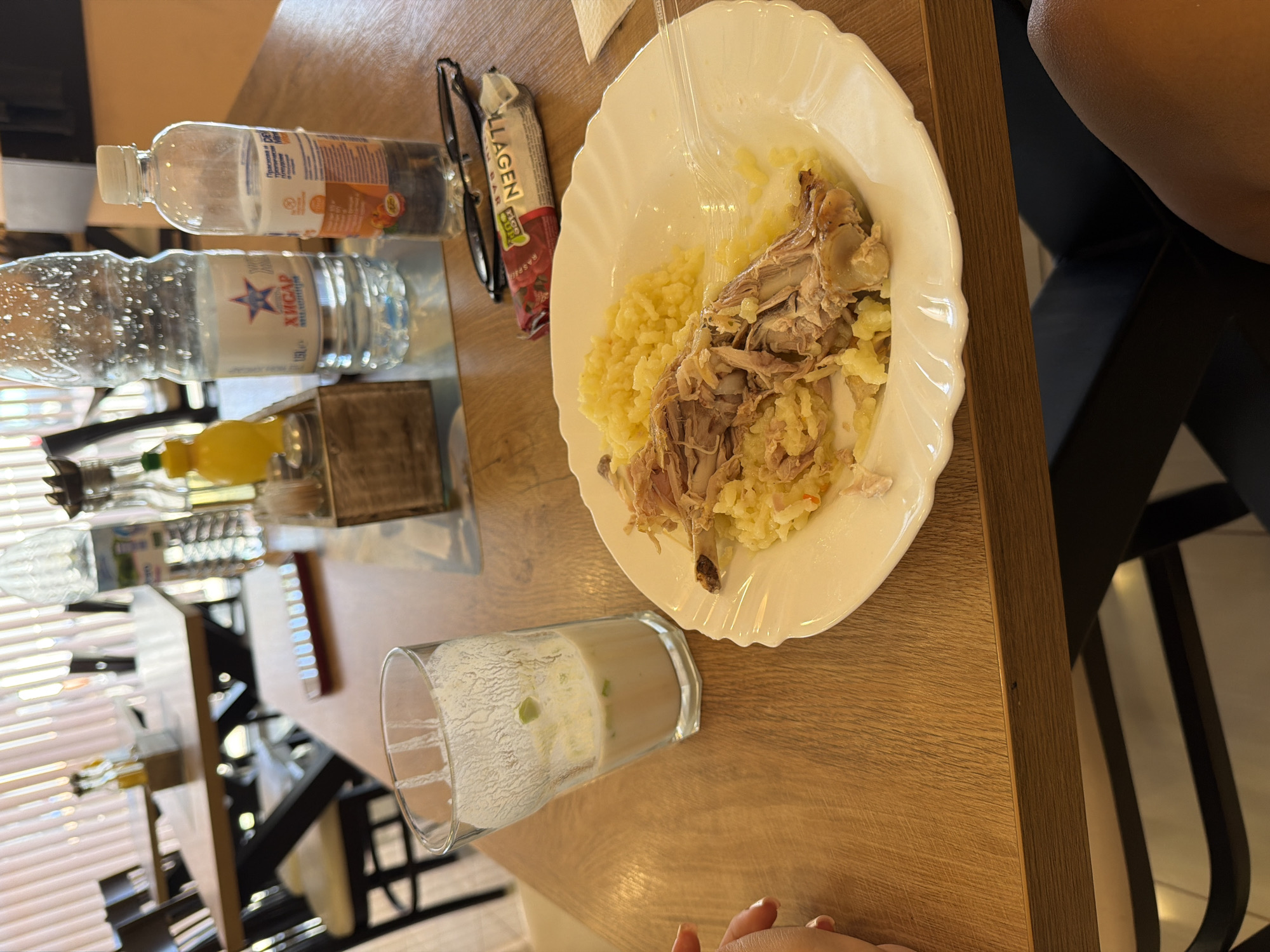 The image shows a table in a restaurant in Burgas, Bulgaria. On the table, there is a plate of food with shredded chicken and mashed potatoes, a glass of drink, a bottle of water, a small packet of a snack, and a pair of eyeglasses. The restaurant appears to be casual, with wooden tables and chairs. The lighting is natural and bright, suggesting it is daytime.