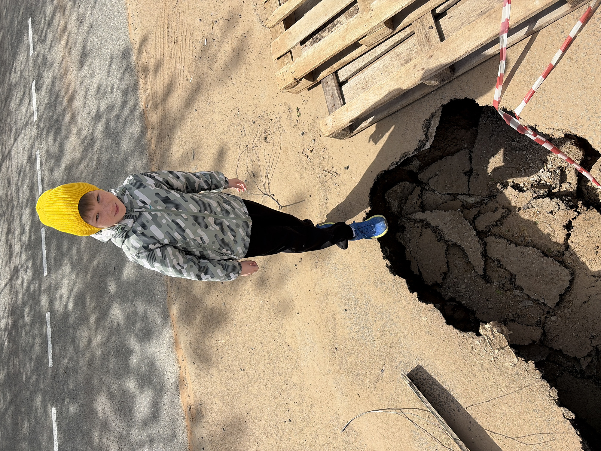 A medium, eye-level shot depicts a young boy in a yellow beanie, a patterned jacket, and blue shoes standing near a large hole in the ground. The hole, surrounded by wooden pallets and red-and-white striped caution tape, appears to be located on a street or sidewalk. The asphalt around the hole is cracked and uneven. The boy is facing the camera with his arms at his sides. The background includes a gray surface with white lines, possibly indicating a road. Shadows from trees or other objects are cast across the gray surface. The location is specified as Unknown, Unknown.