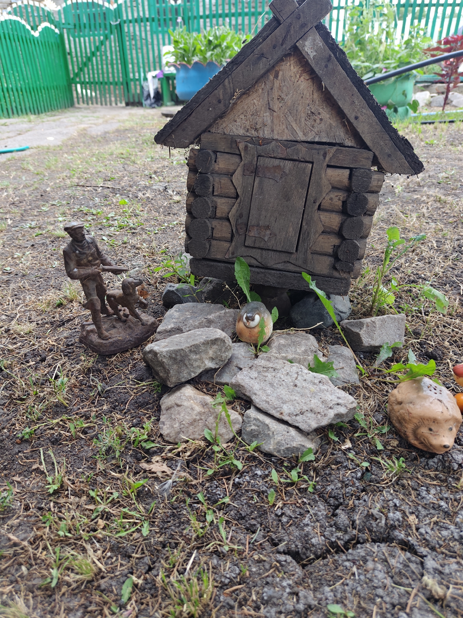 In a rural outdoor setting, possibly in Майкөл ауылдық округі, Kazakhstan, a small, rustic, log-built structure resembling a miniature house stands on the ground. In front of the house, a bronze-colored statuette depicts a man with a hat and a rifle, accompanied by a dog. Several rocks of various sizes are scattered around the scene. To the right of the house, a ceramic figurine of a hedgehog is visible. The ground is covered with dry grass, soil, and some small green plants. In the background, a green picket fence and lush vegetation are present.