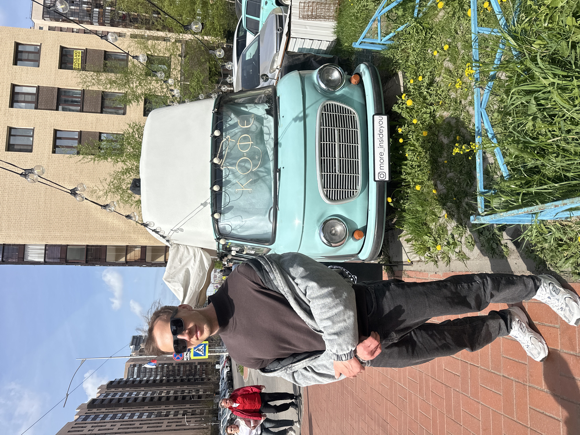 The image is taken in the city of Murino, Russia, showing a man posing in front of a vintage light blue and white mobile coffee truck with the word "Kofe" written in neon lights inside the windshield. The man is wearing sunglasses, a dark brown long-sleeved shirt, a gray jacket, black jeans, and white sneakers. He is standing on a brick sidewalk, with a crosswalk sign visible in the background. Behind him, other people are walking on the sidewalk, including a person in a red jacket. The truck is parked on a patch of grass with yellow dandelions, and a blue metal fence surrounds the area. Behind the truck is a brick building with multiple windows, and there are string lights hanging above the truck.