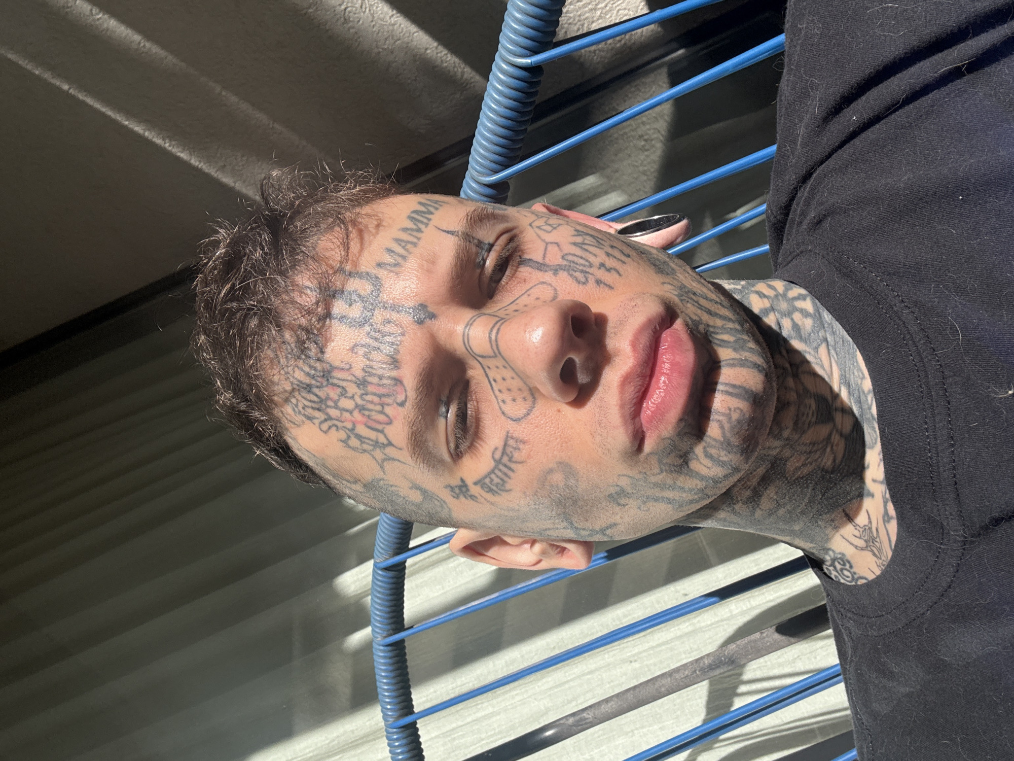 A close-up portrait of a man with extensive face and neck tattoos. He has short, dark hair and light eyes. He is wearing a black t-shirt and has a stretched earlobe visible. He is seated in a blue, tubular chair. The background appears to be a partially shaded, corrugated metal or plastic surface. It is impossible to ascertain the location in the city of Buenos Aires, Argentina without additional information.