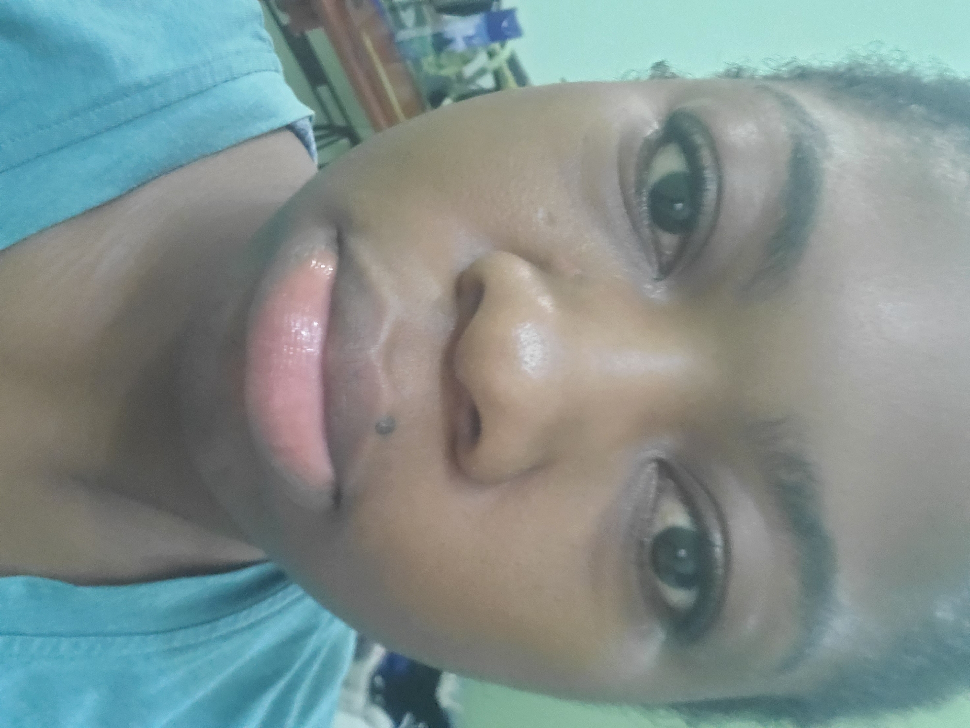 The image is a close-up, frontal portrait of a young Black woman's face. She is looking directly at the camera. Her skin appears to be a medium to dark brown tone, with some slight sheen. Her eyebrows are dark and moderately thick. Her eyes are dark and appear to have eyeliner applied. Her nose is broad. Her lips are full and appear to have a lip gloss or balm on them, giving them a glossy finish. There is a small mole on her upper lip, slightly to the left of center. She is wearing a turquoise-colored t-shirt. The background is out of focus and appears to be an indoor setting with a light green wall and some objects on a shelf or table to the left, including what looks like bottles and a wooden surface. The lighting suggests an artificial light source. There is no indication of the city of Barden Yana, Nigeria, within the image itself.