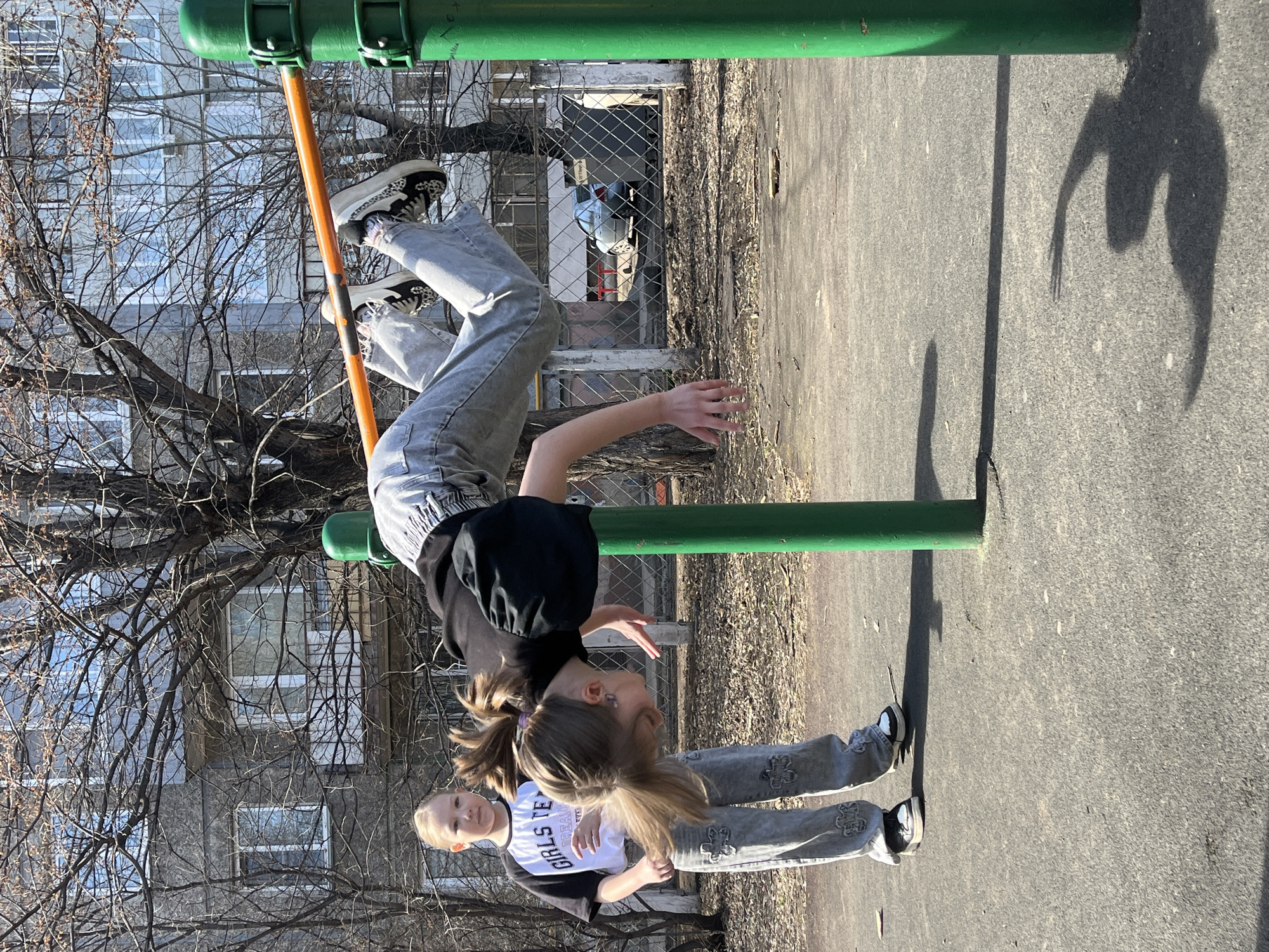 This image, taken in Krasnoyarsk, Russia, depicts two young females in an outdoor urban setting, likely a playground or park. The scene is bathed in bright sunlight, casting long, distinct shadows on the asphalt ground, suggesting it is late morning or afternoon.
One girl, with light brown hair styled in pigtails, is actively engaged in acrobatics. She hangs upside down from a green horizontal bar, suspended by her legs, with her arms outstretched. She is dressed in a black, puff-sleeved top and light-wash jeans, complemented by black and white patterned sneakers.
Standing to her right and slightly behind is another girl with blonde hair, observing the first. She wears a white t-shirt over a black long-sleeved shirt, light-wash jeans with embroidered or patched details, and black and white sneakers. The text "GIRLS TEAM TREAT" is visible on her white t-shirt.
An orange vertical pole supports the green horizontal bar. In the background, a multi-story residential building with numerous windows is visible through the sparse branches of bare trees, indicating either late winter or early spring. A chain-link fence is also present in the middle ground. The image captures a moment of playful activity.