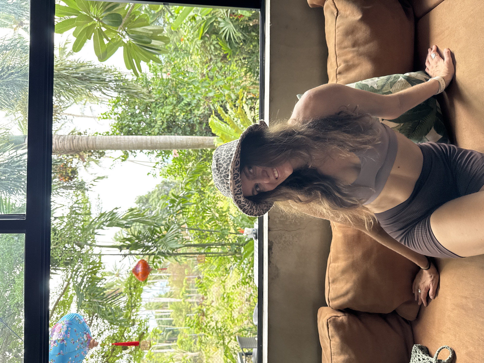 A woman wearing a leopard print bucket hat, a sports bra, and athletic shorts sits on a brown couch. She looks at the camera and smiles. Outside the window is dense, green foliage, including palm trees. The sky is overcast. In the foliage are hanging decorations: a red balloon, and a blue flowered umbrella. The location is likely Baan Mae Nam, Thailand, as indicated in the prompt.