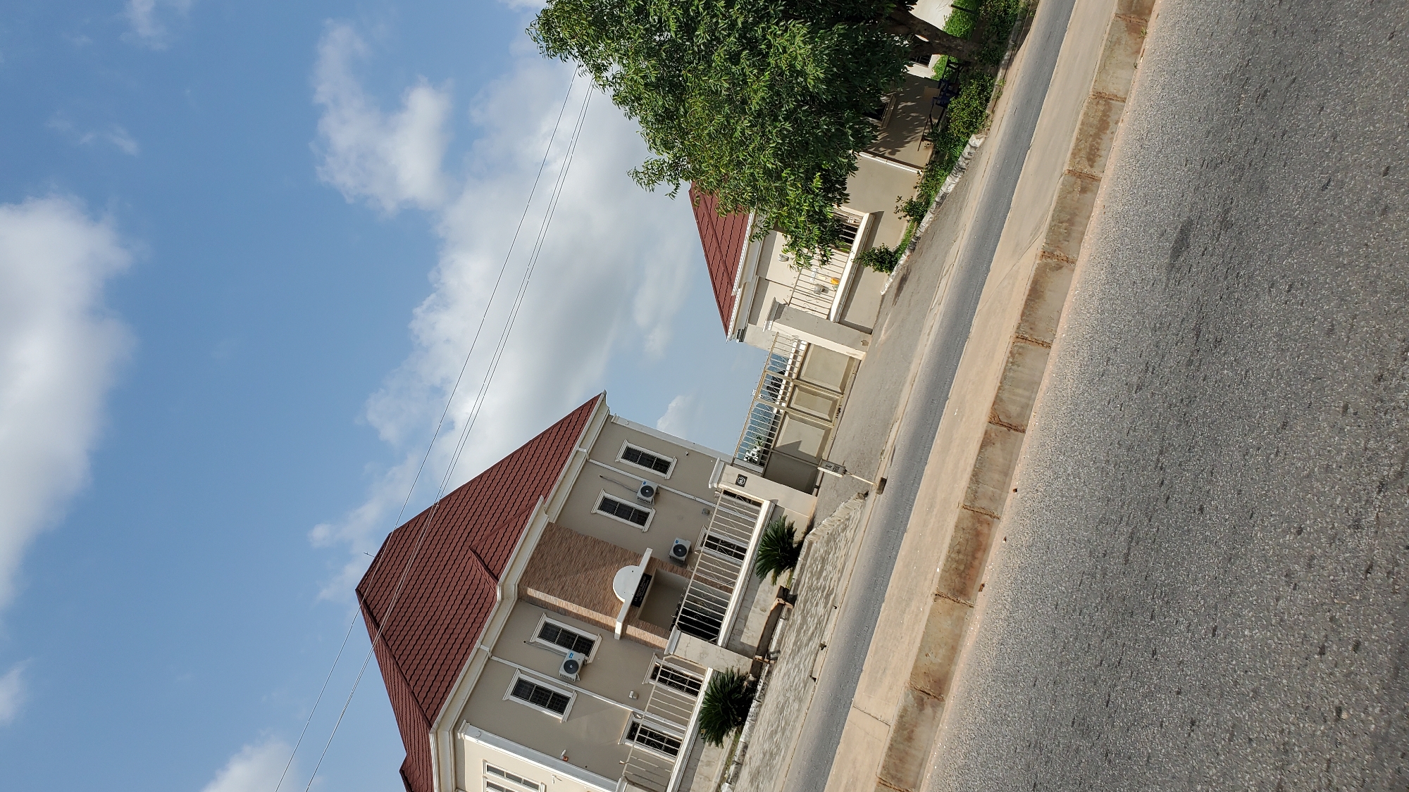 The media file depicts a street scene in Jalingo, Nigeria, featuring two modern residential buildings with red tiled roofs and light-colored facades. The buildings are multi-story, with visible windows and air conditioning units. One building has a stone-accented section on its upper floor. Both properties are enclosed by white metal railings and have small, well-maintained plants in front of them. A beige metal gate is visible on the right side of the scene, leading into one of the properties. A large green tree with dense foliage stands behind and to the right of the second building. The foreground shows an asphalt road with a concrete curb and a drainage channel. The sky is a clear blue with scattered white clouds, suggesting it is daytime with fair weather. Power lines are visible stretching across the sky. There is no visible text in the media file. The overall impression is one of a developed residential area.