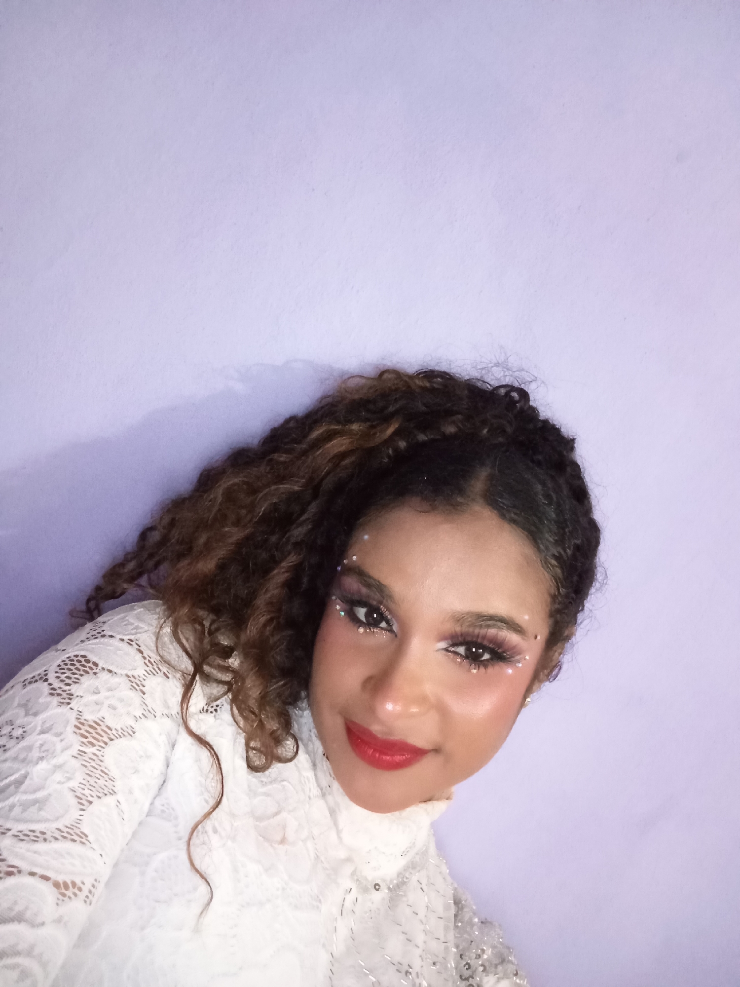 A young woman with curly brown hair and light brown skin is pictured from a low angle against a light purple wall. She is wearing a white lace top with a high collar. Her makeup includes red lipstick, heavy blush, decorated with several small white gems on her eyebrows and around her eyes. Her gaze is directed at the viewer with a slight smile. The background is plain and featureless. There is no information provided to determine the location of this image in the city of Juan Griego, Venezuela.