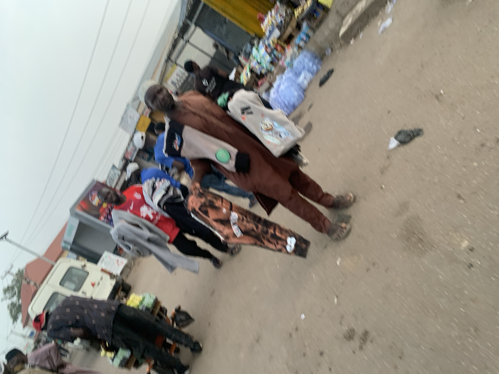 This image captures a busy street market scene in Jos, Nigeria, under an overcast sky, suggesting a cloudy day with diffuse lighting. The perspective is slightly elevated and angled, looking down a thoroughfare bustling with people and merchandise.
In the foreground, a man wearing a brown, possibly traditional, long-sleeved outfit walks towards the right side of the frame. He carries a light-colored bag on his shoulder that has "LUCKY BEAM" and cartoon-like graphics printed on it, and another item with a green circular patch. A patterned brown garment, possibly for sale, is laid out horizontally in the mid-ground.
Several other individuals are visible throughout the market. To the left, a man in a patterned dark shirt and dark pants stands near a white van. Further into the scene, other people are seen, some wearing red shirts, others in blue or dark clothing, appearing to be engaged in shopping or selling activities. Merchandise, including clothes, packaged goods, and bottles, is displayed on makeshift stalls and tables along the edges of the street.
The ground is a light grey or beige color, possibly concrete or packed earth, with scattered debris such as small papers and abandoned footwear (a black sandal and a grey slipper). In the background, simple buildings, some with yellow facades or corrugated metal roofs, line the street, indicating a commercial area. The overall scene depicts a typical, active market environment. Visible text includes "LUCKY BEAM" on the man's bag, and blurry text on other garments and signs in the background.