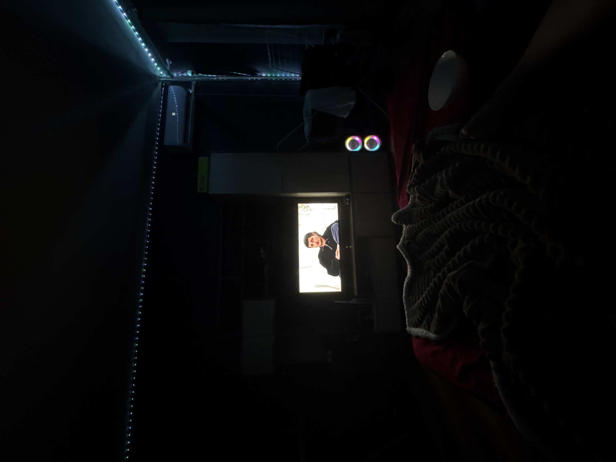 A dimly lit room is shown, rotated ninety degrees counterclockwise. A person is visible in a brightly lit doorway. The room contains LED strip lights along the ceiling and a vertical strip along one wall. Other items include a drying rack, a white container, two illuminated orbs, a cup, and a textured blanket on a red surface. The location is not discernible from the image.