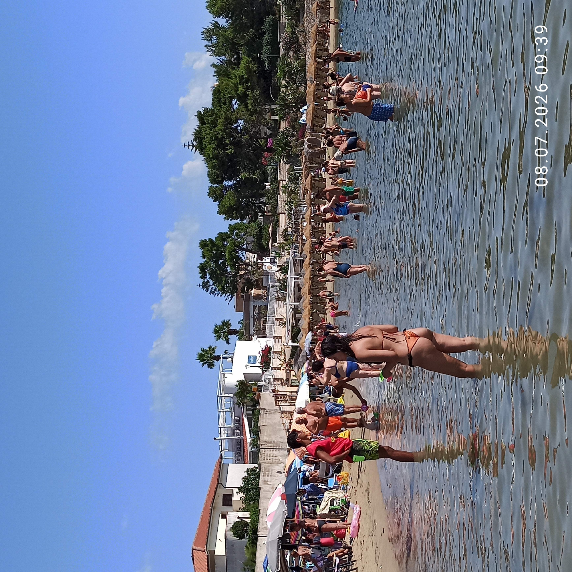 The image depicts a crowded beach scene in Trani, Italy, with numerous people wading in the shallow water or standing on the sandy shore. In the foreground, several individuals are visible in the water, some appearing to interact or play. A woman in an orange bikini is in the center foreground, facing away from the camera. The beach is lined with numerous umbrellas and lounge chairs, indicating a popular tourist destination. Buildings, some with balconies and white railings, are visible in the background, along with dense green foliage, including palm trees, suggesting a warm, sunny climate. The water is calm and reflects the sky, which is a clear blue with scattered white clouds. The timestamp "08.07.2026 09:39" is visible in the bottom right corner of the image.
