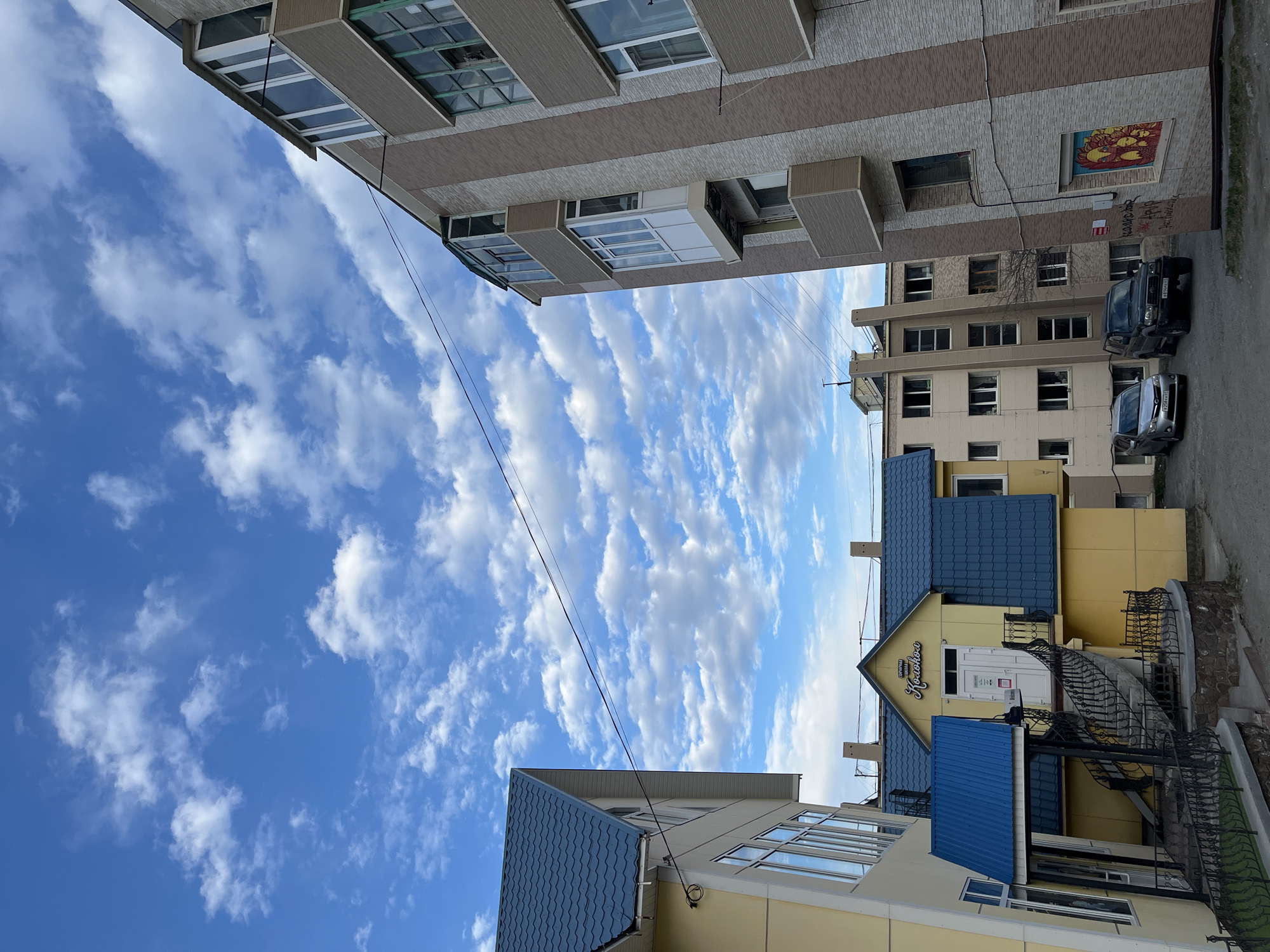 The image depicts a street view in Petropavlovsk-Kamchatsky, Russia, characterized by a mix of architectural styles under a bright, partly cloudy sky. The buildings are varied in height and design. One building has light brown brick with a decorative mural on one side. Two vehicles, a dark SUV and a light-colored sedan, are parked beside it. Another building features light yellow cladding with a blue tiled roof and a black wrought-iron staircase leading to the entrance, which displays the word "Krasnova" in Cyrillic script. Overhead power lines stretch across the sky.